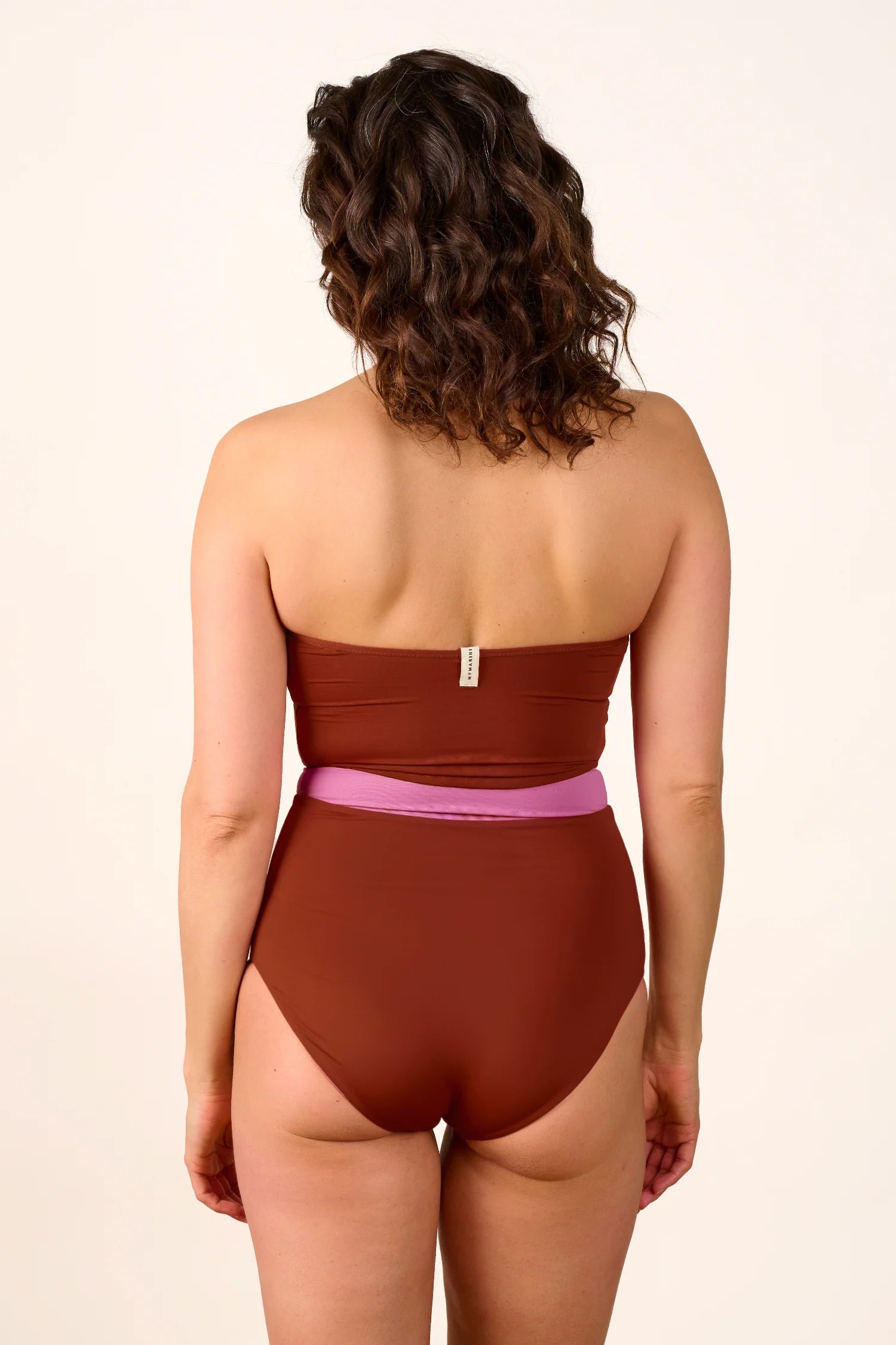 Rückansicht einer Frau in einem MYMARINI Badeanzug mit Bandeau-Schnitt und hohem Beinausschnitt in Braun und Rosa.#color_sienna-orchid