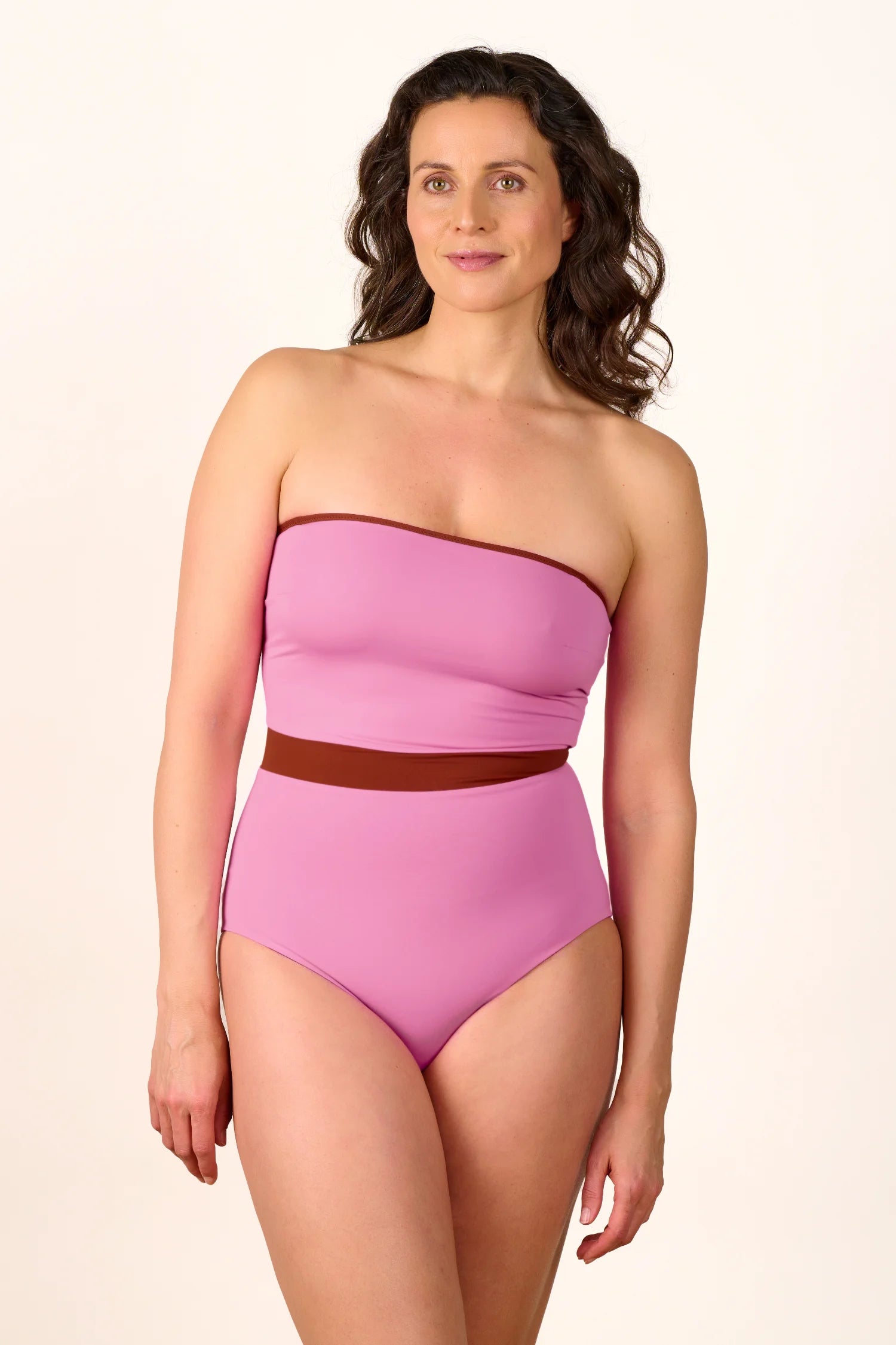 Frau trägt MYMARINI One-Piece in Rosa, Bandeau-Schnitt, hoher Beinausschnitt, vor neutralem Hintergrund.#color_sienna-orchid