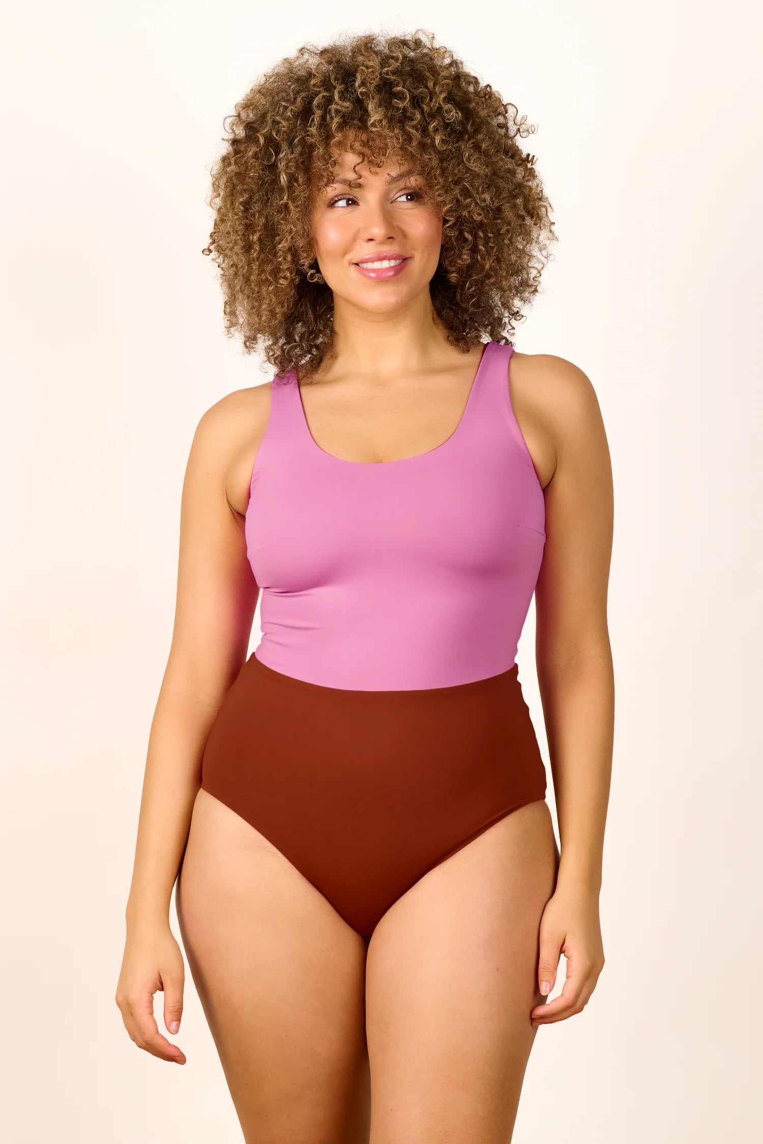 Frau trägt MYMARINI Badeanzug mit breiten Trägern und hohem Beinausschnitt in Rosa und Braun, lächelnd vor neutralem Hintergrund.#color_sienna-orchid