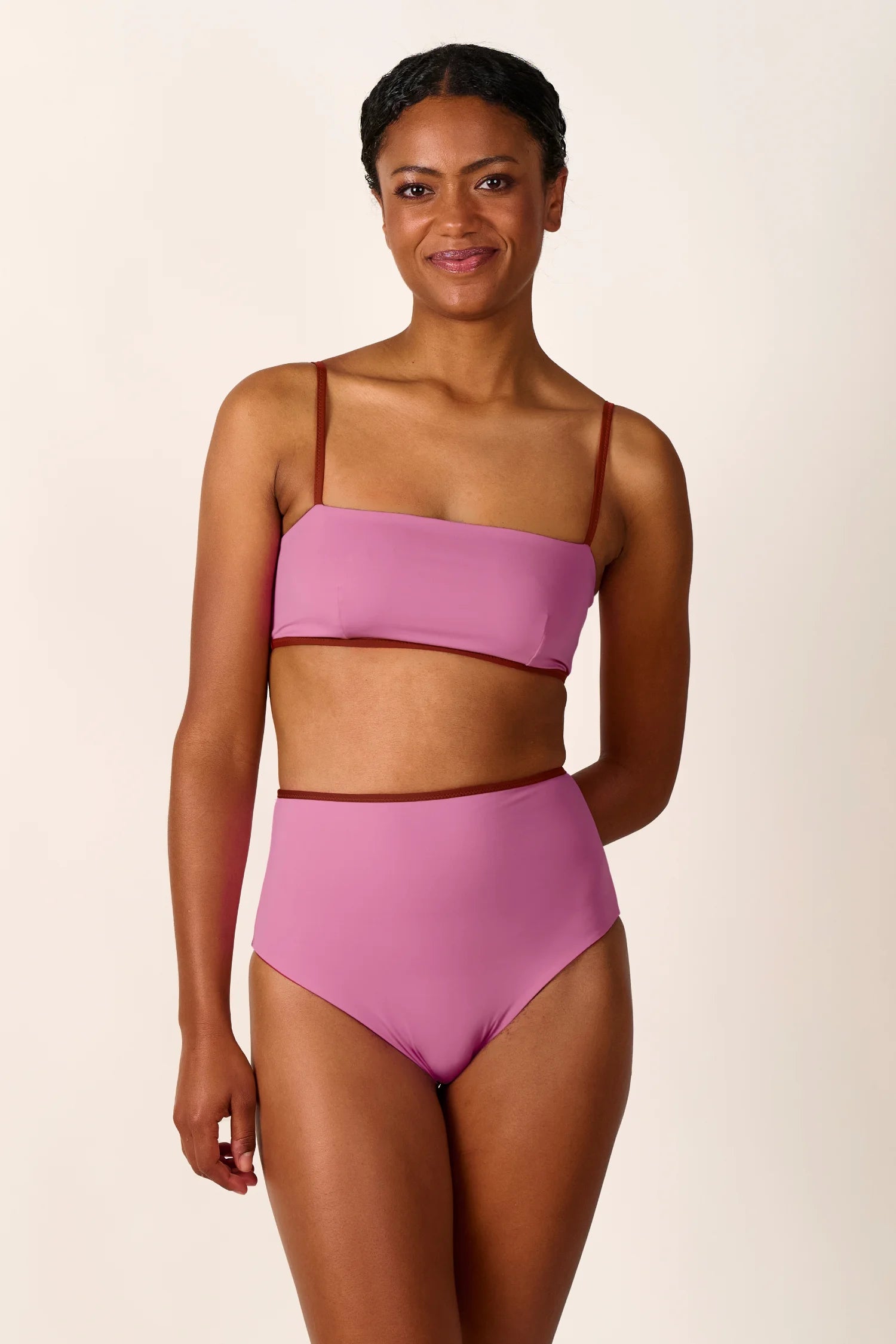 Frau trägt MYMARINI Bikini mit Bandeau-Top und High-Waist-Hose in Rosa, lächelnd vor neutralem Hintergrund.#color_sienna-orchid