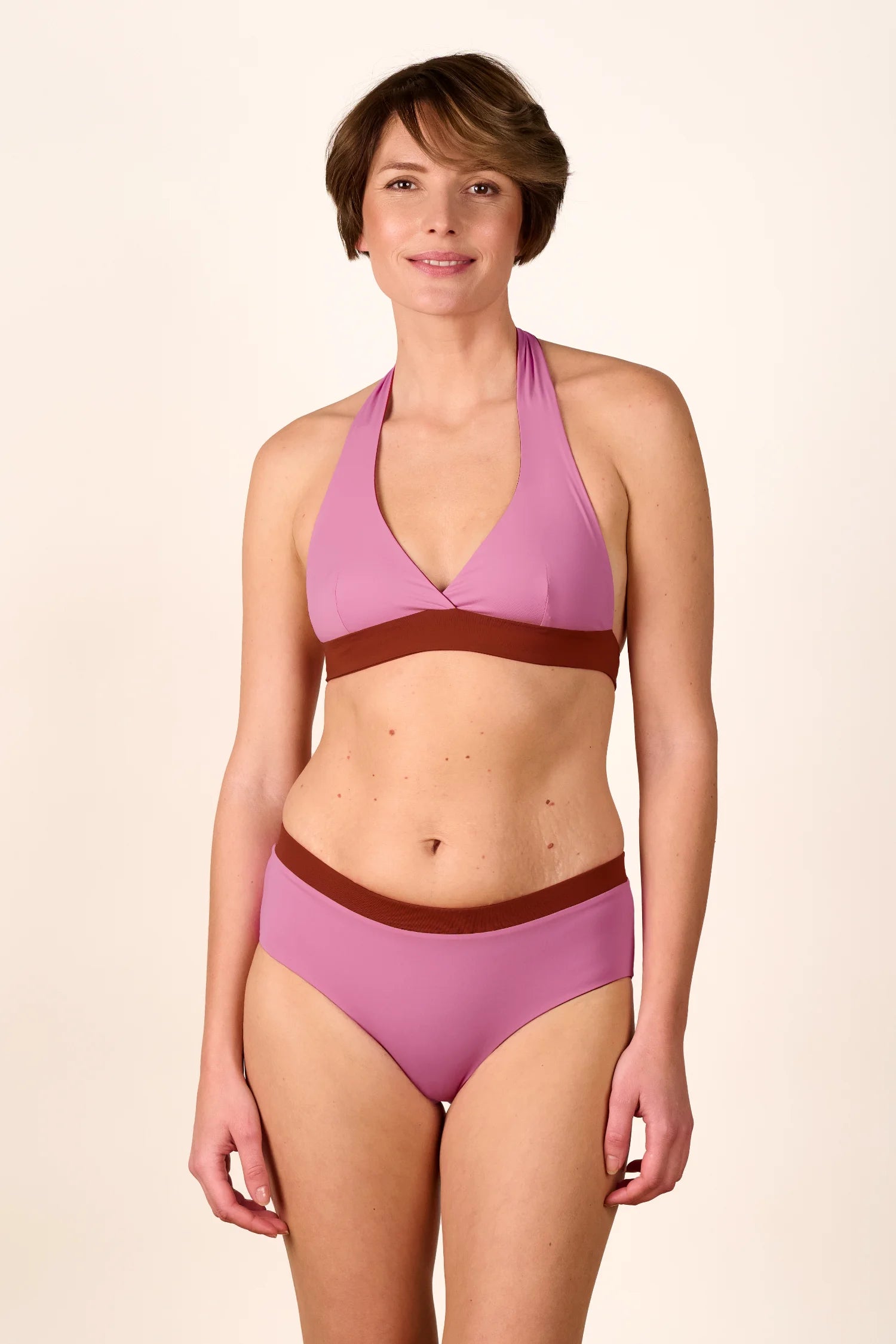 Frau trägt MYMARINI Bikini mit Neckholder-Top und High-Waist-Hose in Rosa und Braun, lächelnd vor neutralem Hintergrund.#color_sienna-orchid