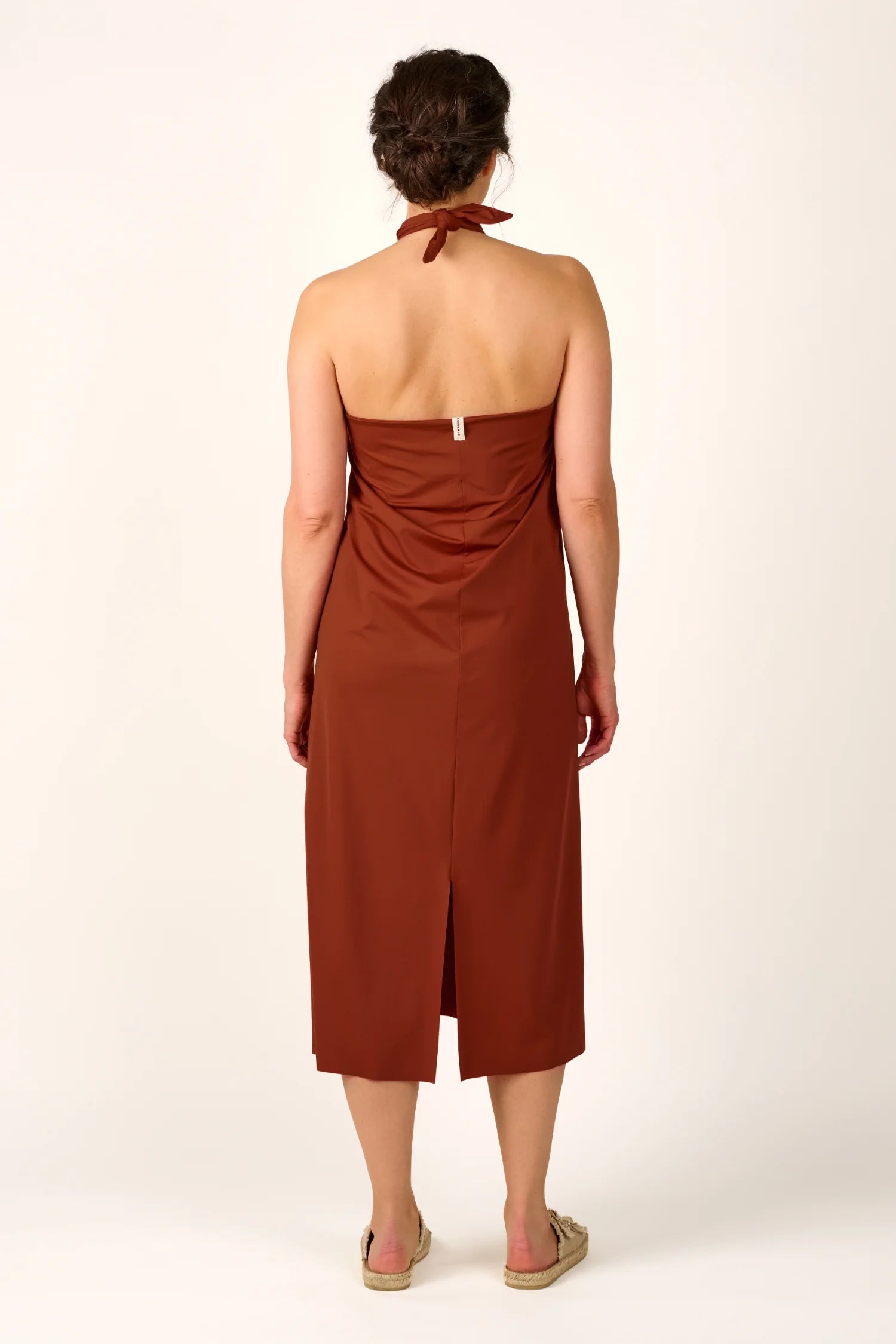 Person von hinten in einem braunen MYMARINI Neckholder-Kleid mit mittellangem Schnitt und Schlitz, barfuß auf hellem Hintergrund.#color_sienna