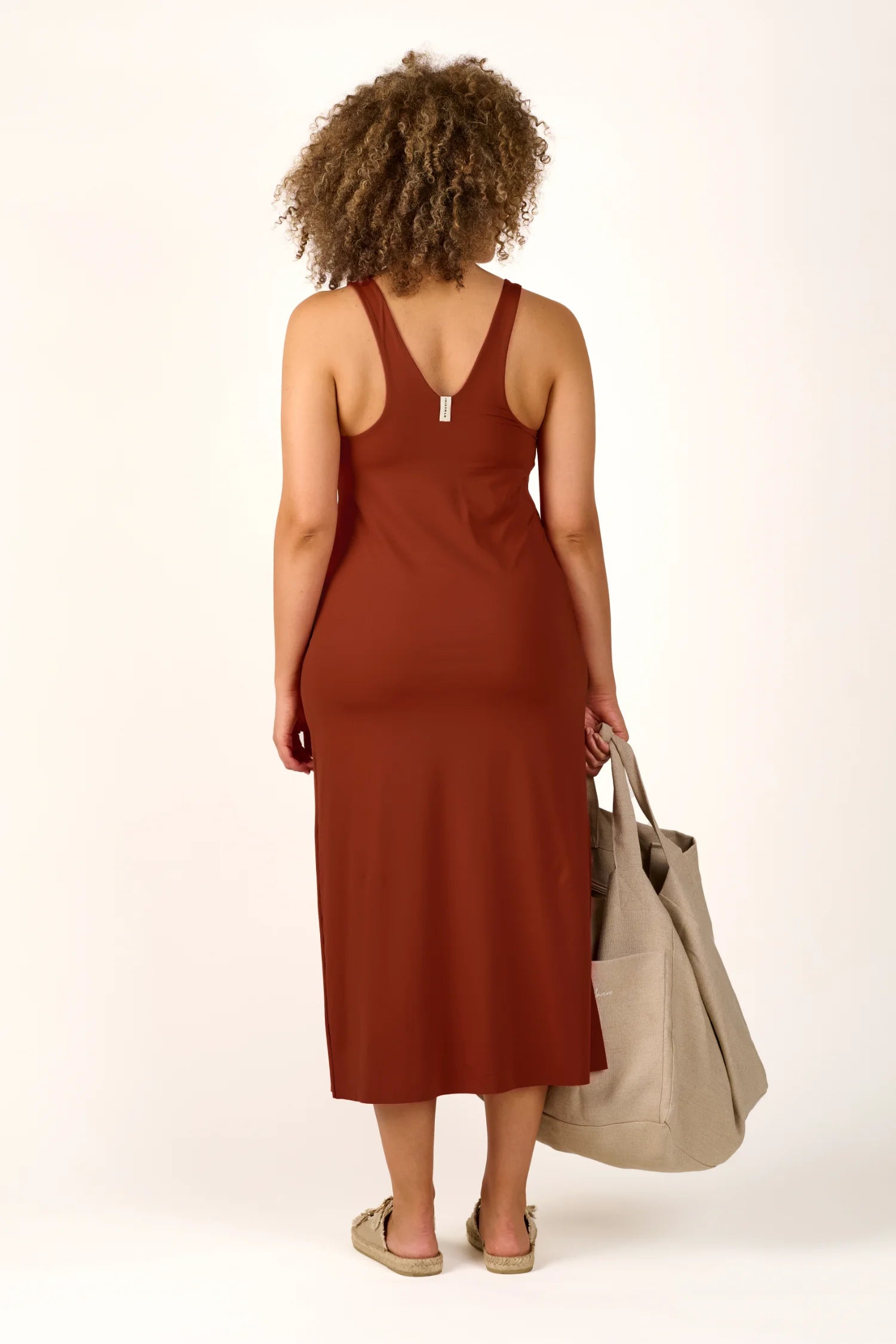 Person von hinten in einem ärmellosen, hoch geschlossenen MYMARINI-Kleid, hält eine große Tasche.#color_sienna