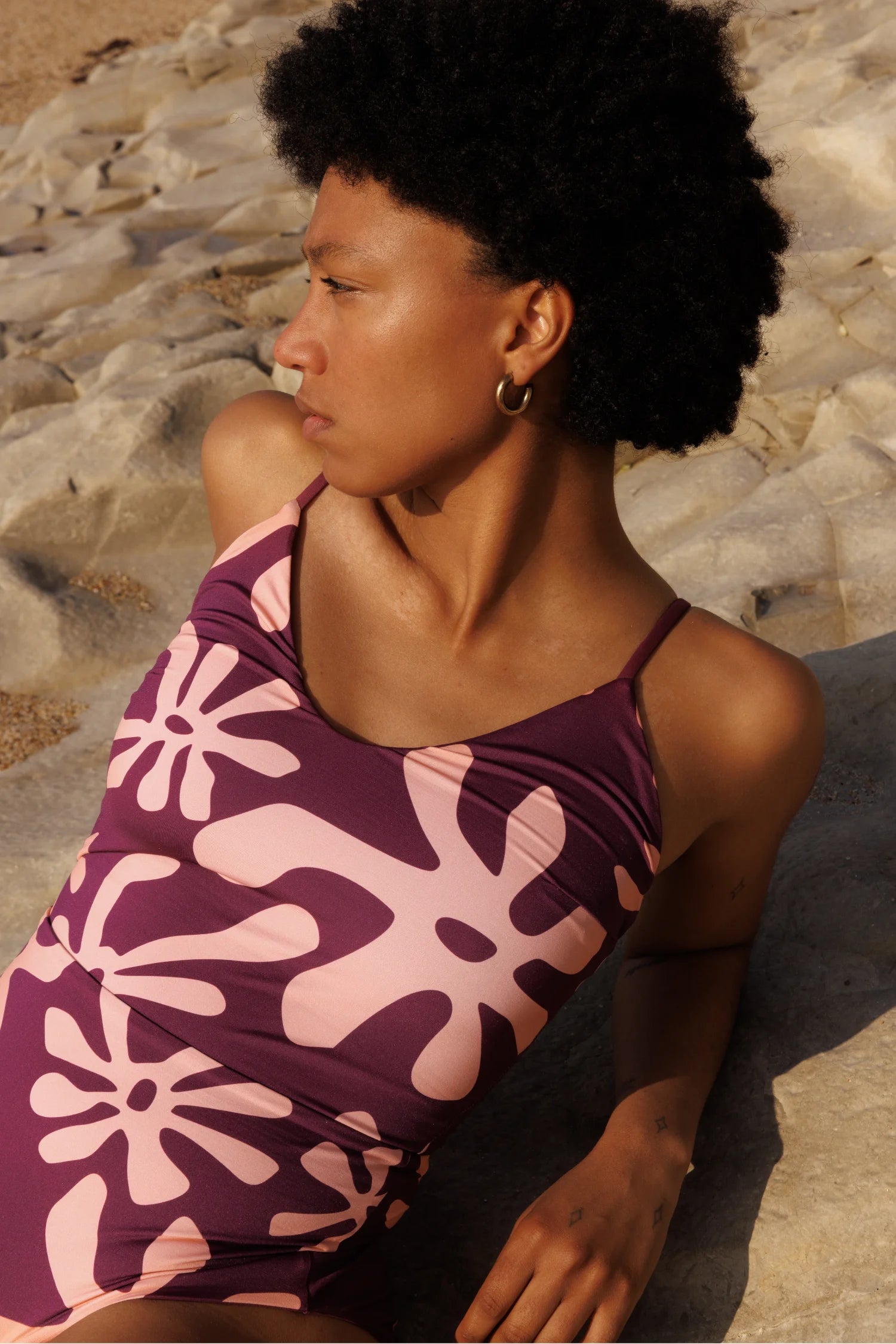 Badeanzug von MYMARINI in lila mit pinkem Muster, hochgeschlossen, getragen von einer Person auf Steinen am Strand.#color_berry-print