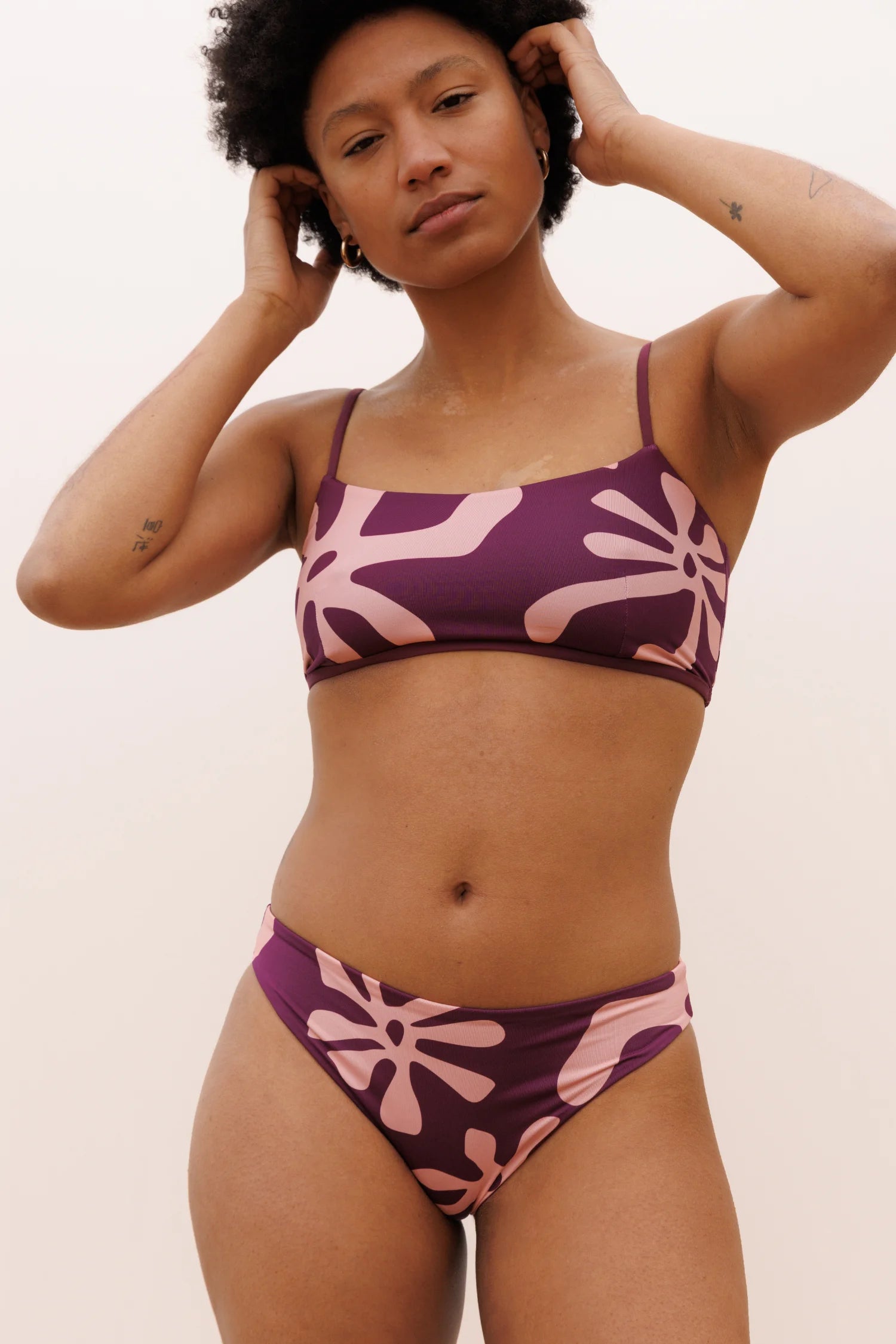 Bikini von MYMARINI in lila mit pinken Mustern, Spaghettiträger und tiefer Beinausschnitt. Model posiert mit den Händen im Haar.#color_berry-print