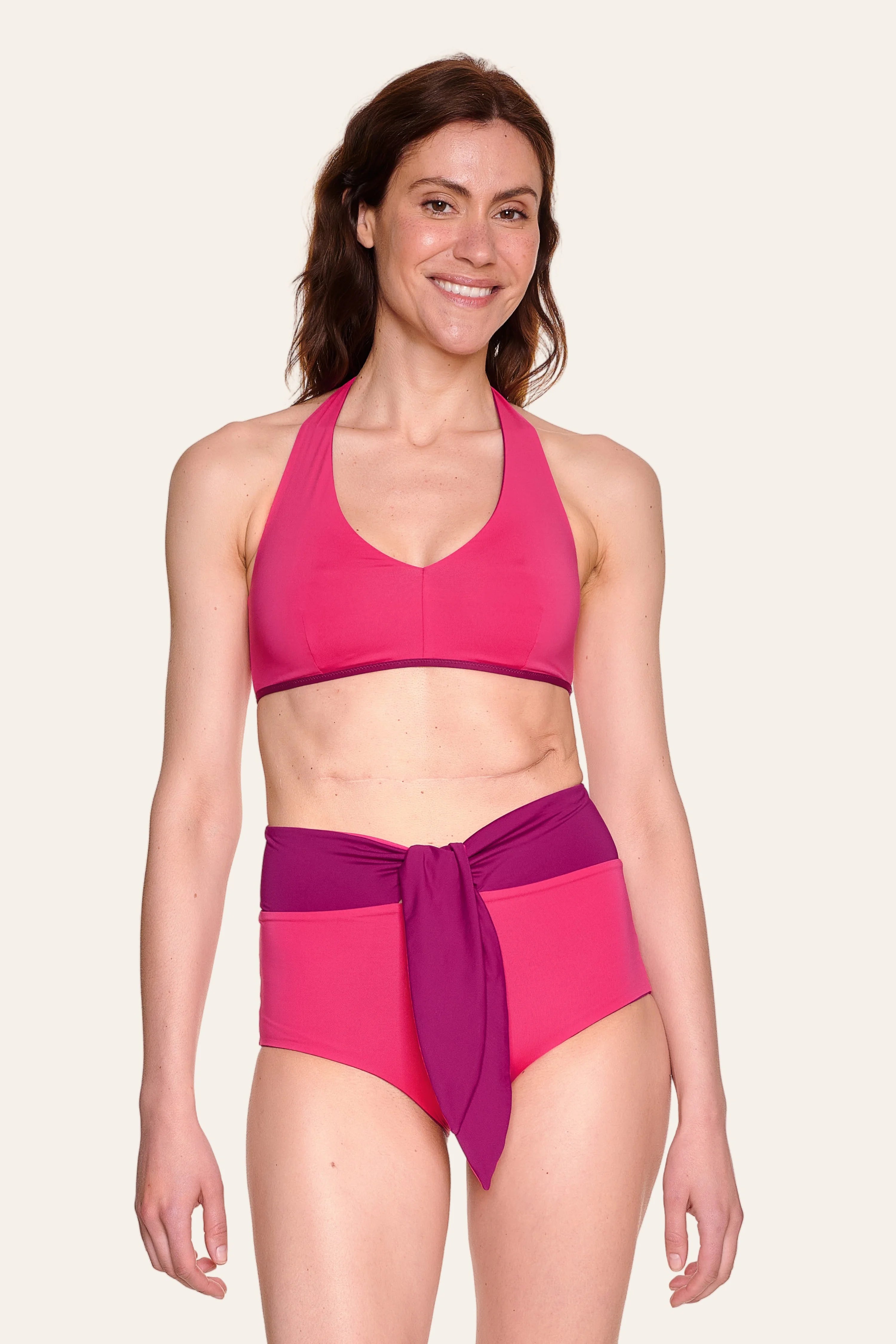 Frau trägt MYMARINI Bikini mit Neckholder-Top und High-Waist-Hose in Pink und Lila, lächelnd vor hellem Hintergrund.#color_pink-hortensia