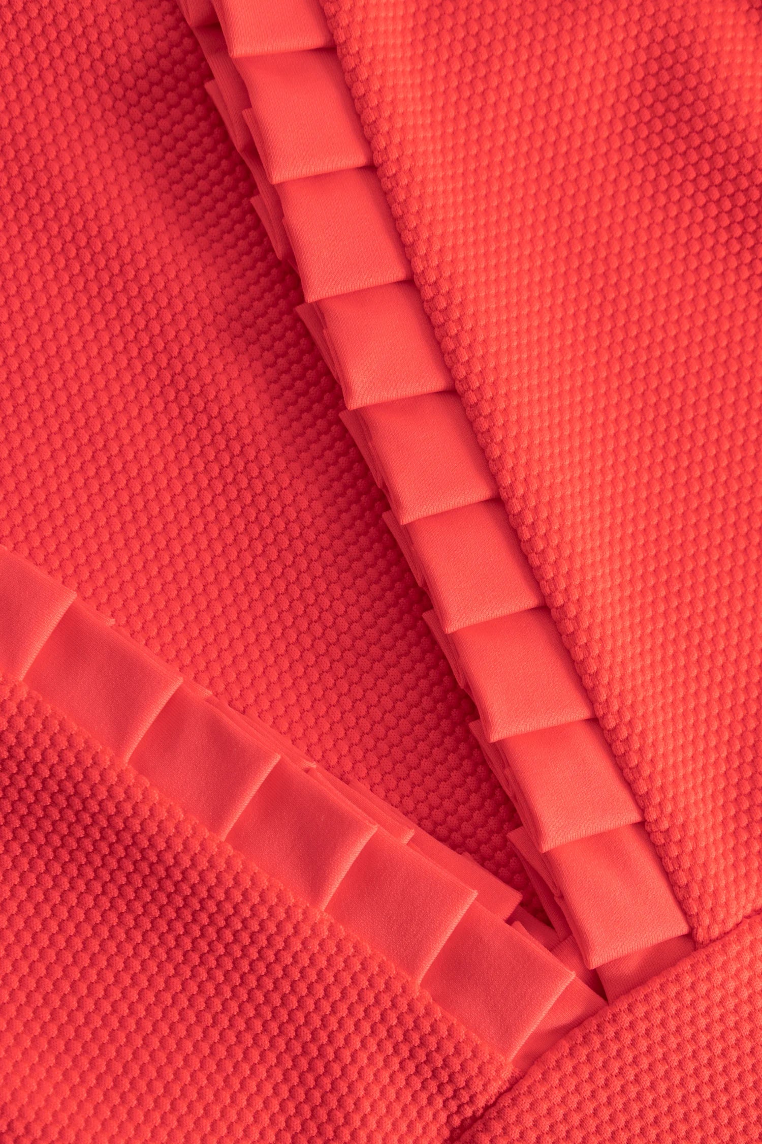 Nahaufnahme eines orangefarbenen, strukturierten Bademodestoffs mit geriffelter Oberfläche und umlaufenden Faltkanten.#color_rouge