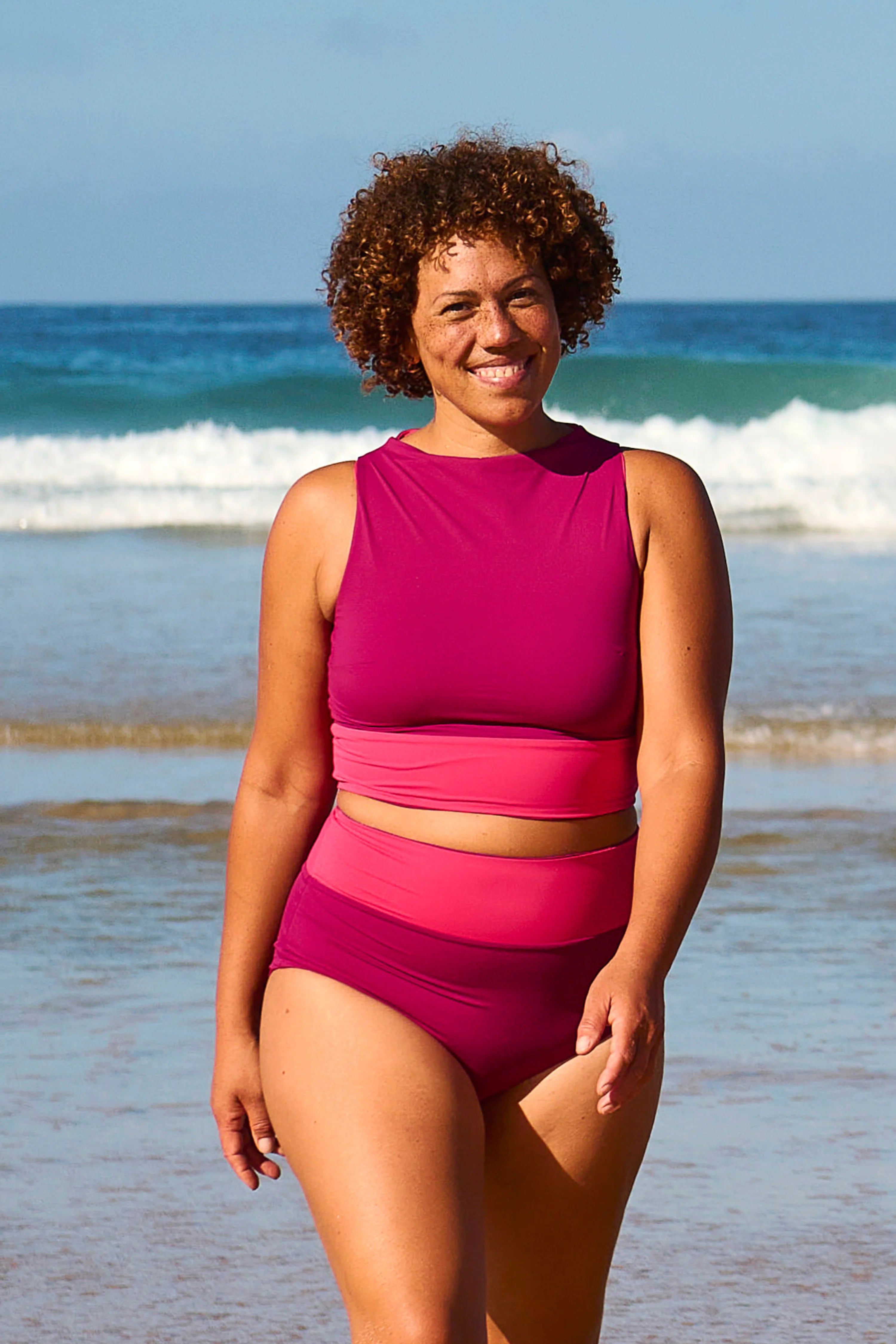 Frau am Strand in MYMARINI Bikinihose und Top, hoch geschlossen, mit breiten Trägern, vor Wellen im Hintergrund.#color_pink-hortensia