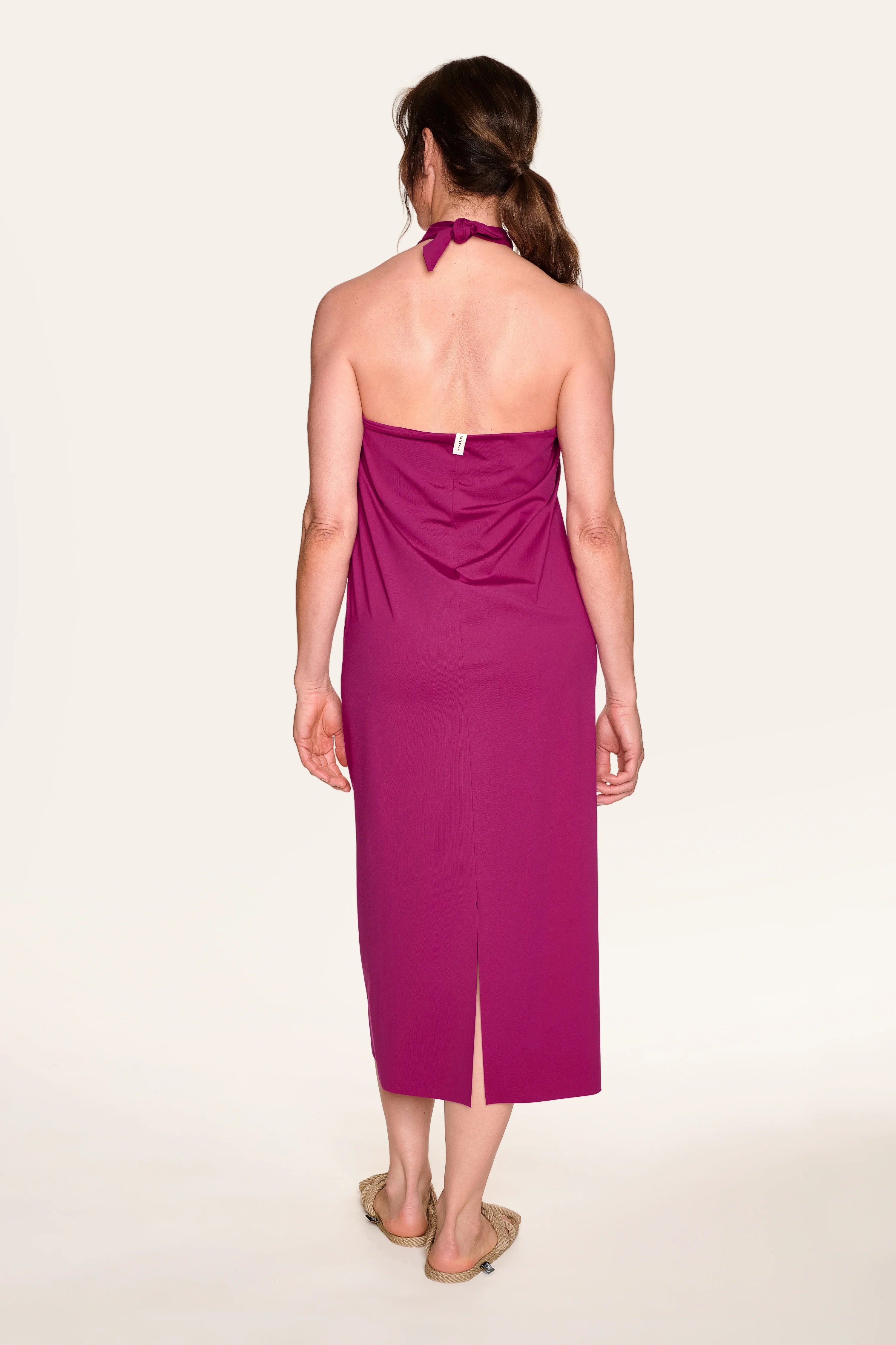 Person von hinten in einem magentafarbenen MYMARINI Neckholder-Kleid mit Schlitz, barfuß auf hellem Hintergrund.#color_hortensia