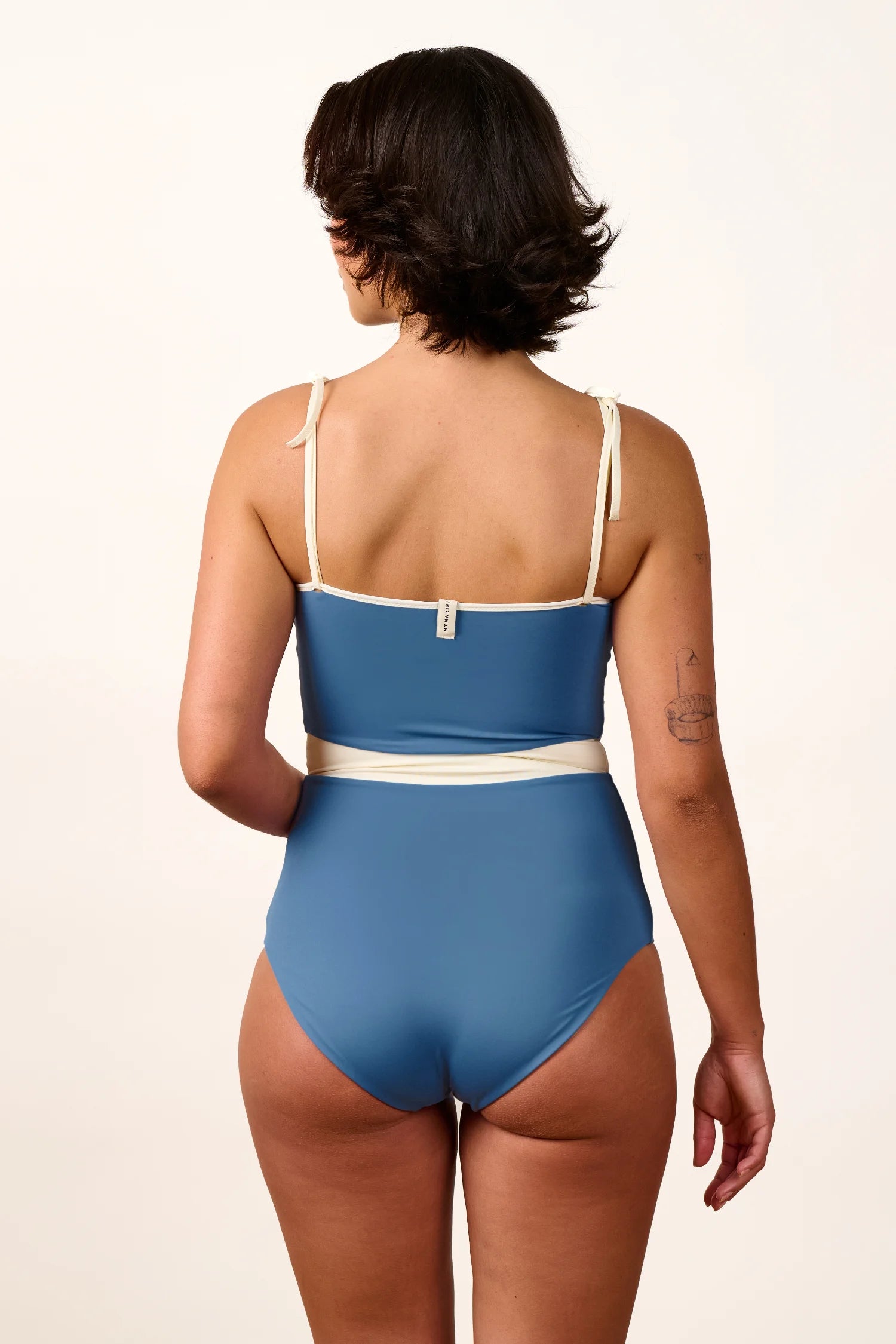 Person in blauem MYMARINI-Badeanzug mit Spaghettiträgern und hohem Beinausschnitt, von hinten gezeigt.#color_coast-desert