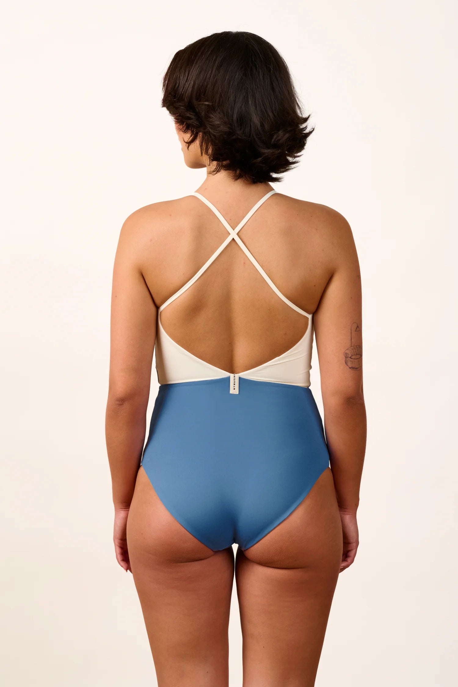 Person trägt einen MYMARINI One-Piece mit hohem Beinausschnitt und Cross-Top-Design in Blau und Weiß, von hinten gesehen.#color_coast-desert
