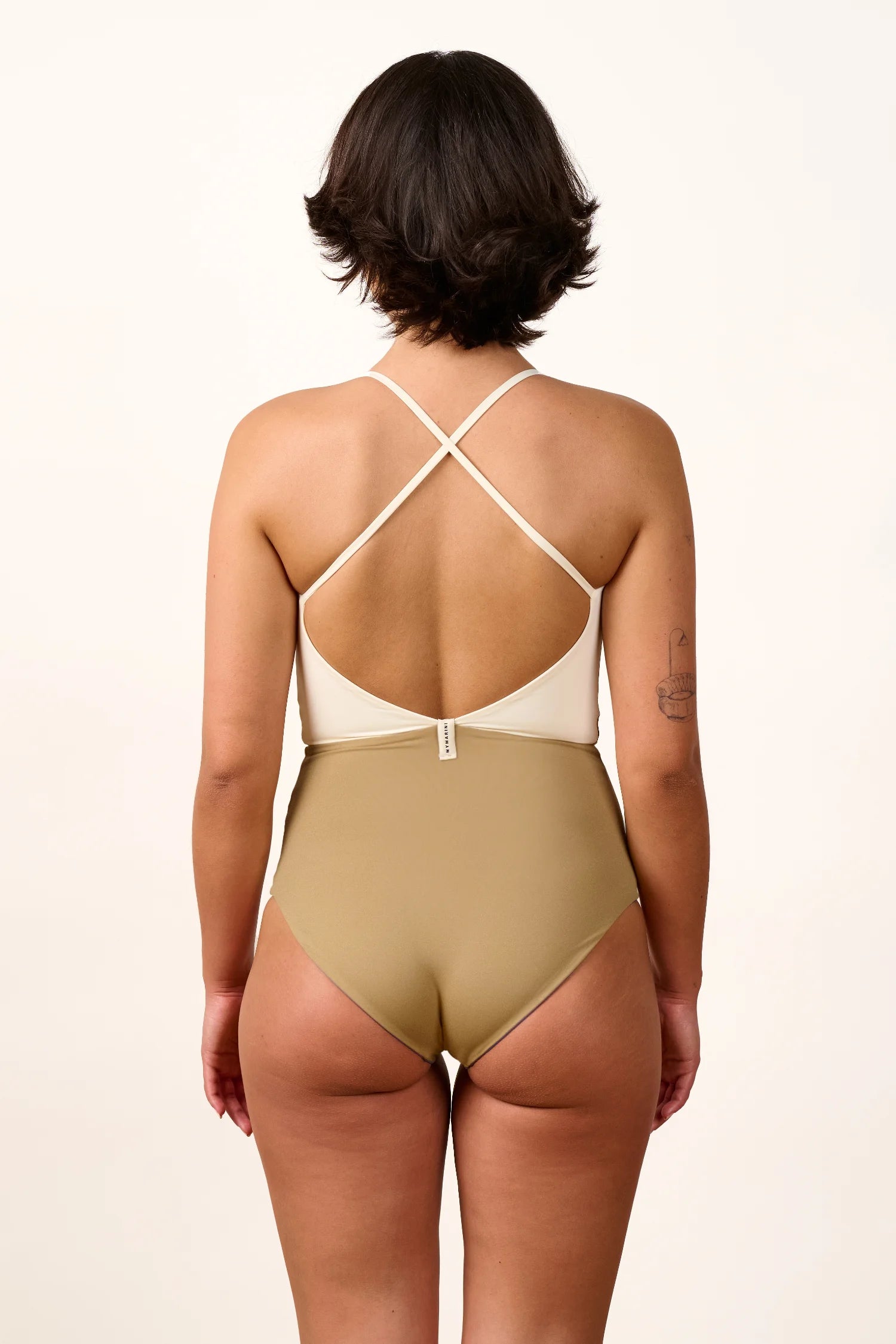 Person von hinten in einem MYMARINI One-Piece mit hohem Beinausschnitt und Cross-Top-Trägern in Beige und Weiß.#color_coast-desert