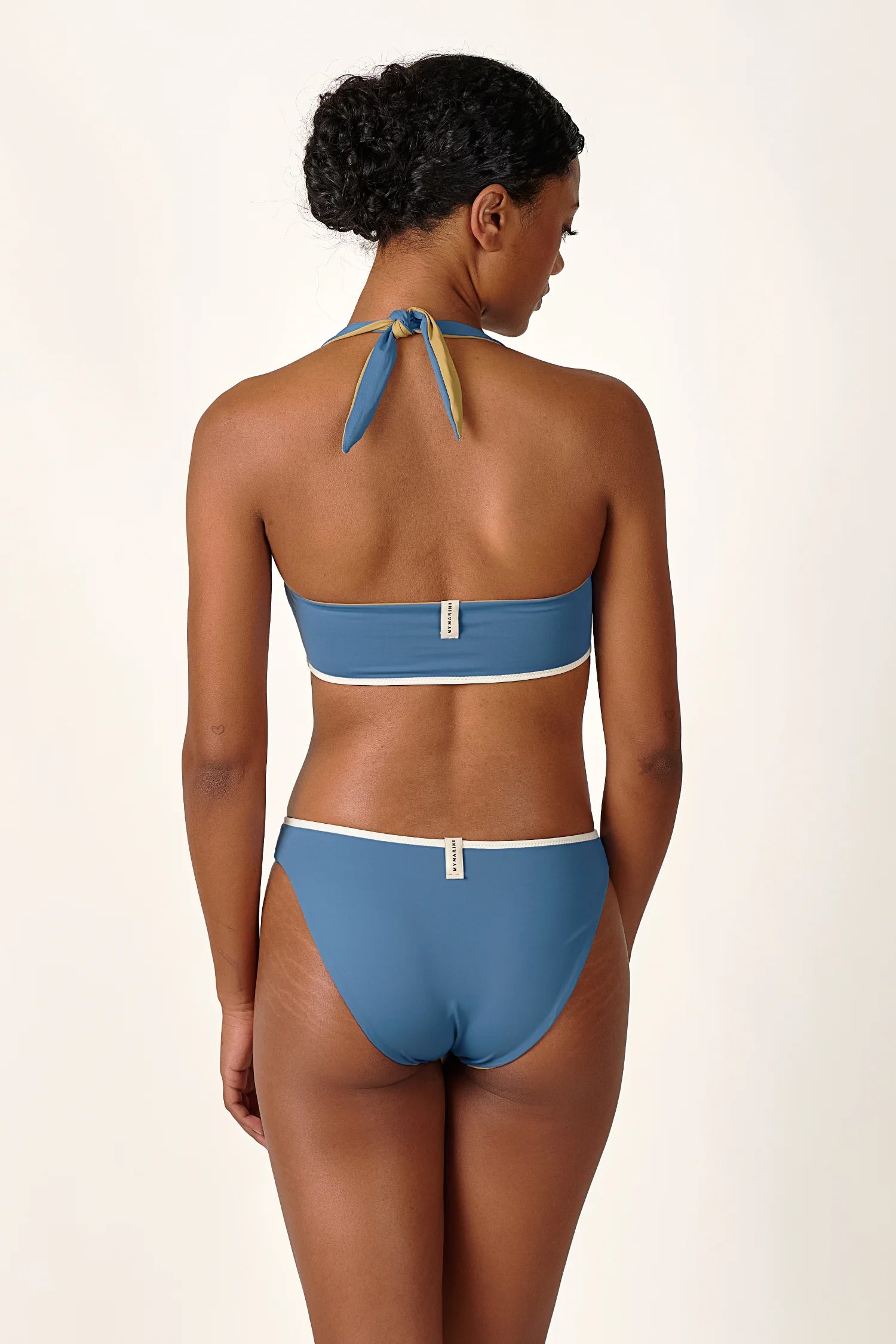 Frau trägt MYMARINI Bikini mit Bandeau-Top und hoher Bikinihose in Blau, Rückansicht, vor neutralem Hintergrund.#color_coast-desert