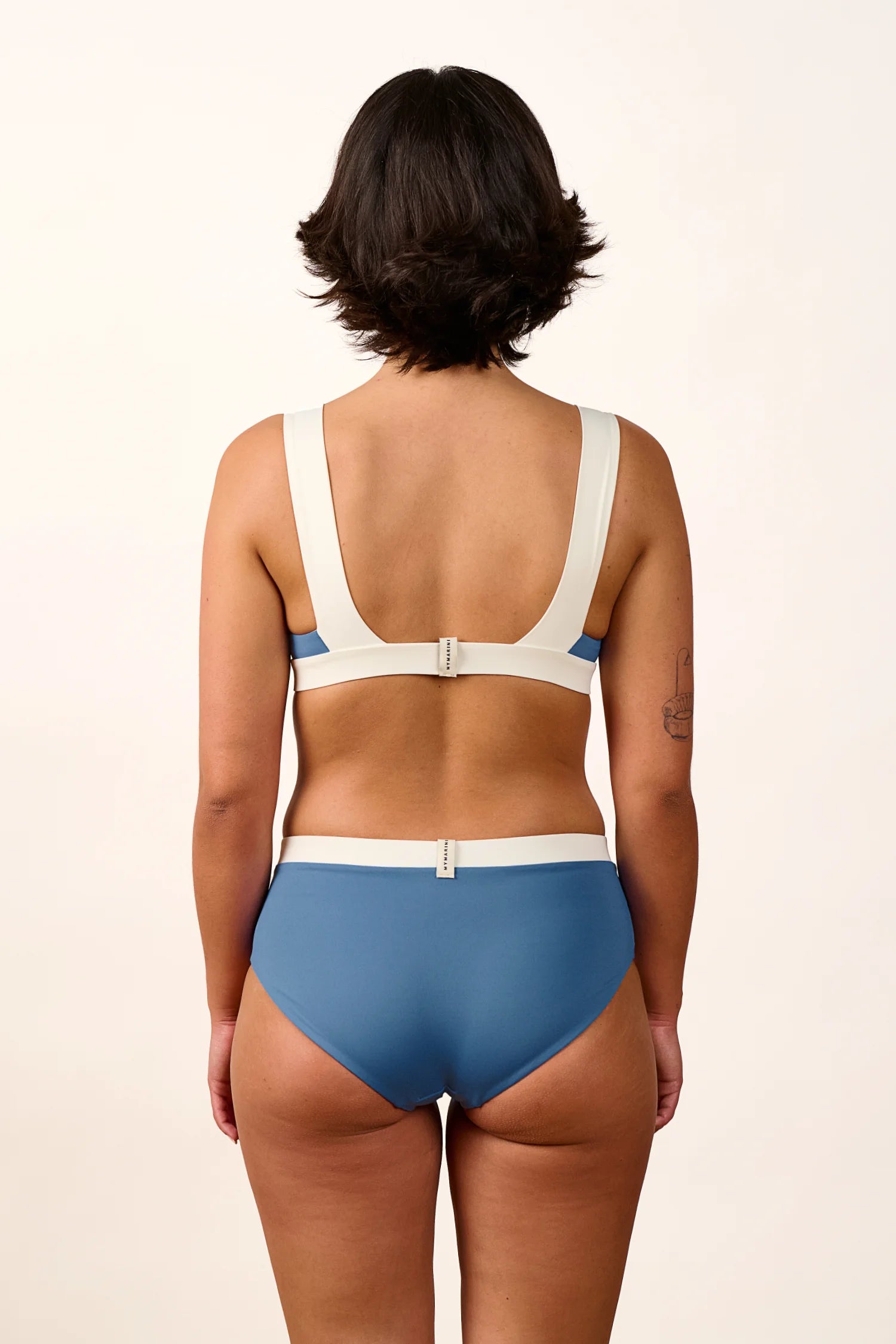Rückansicht einer Person in einem MYMARINI Bikini mit breiten Trägern und hohem Beinausschnitt in Blau und Weiß.#color_coast-desert