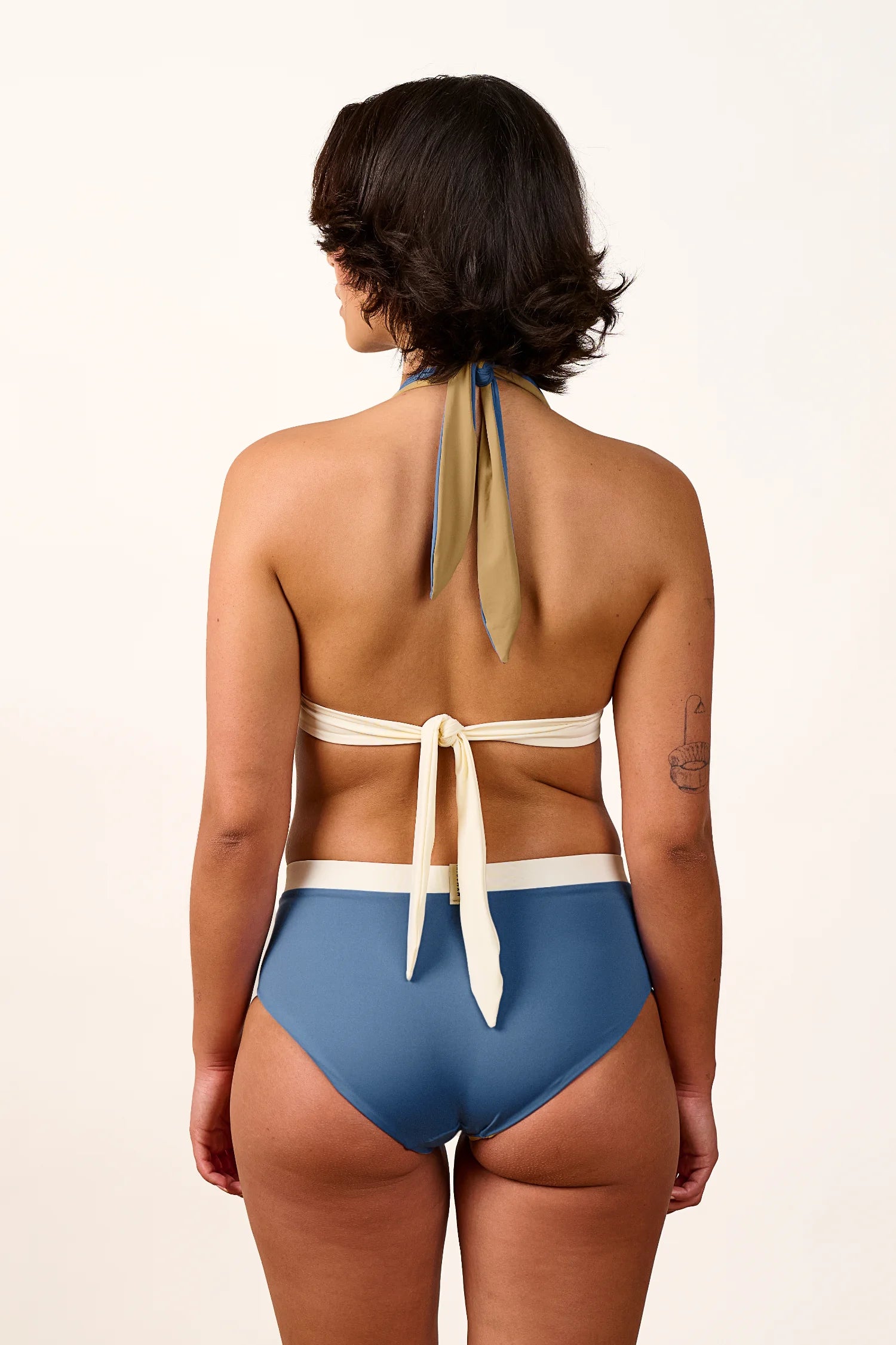 Frau in MYMARINI Bikinihose mit hohem Beinausschnitt und Neckholder-Top, Rückansicht, vor neutralem Hintergrund.#color_coast-desert