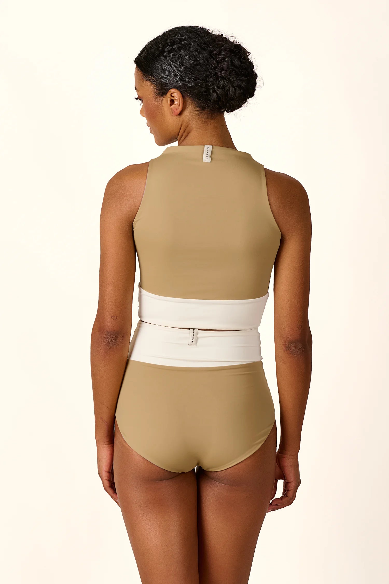 Frau in MYMARINI Badeanzug mit hohem Beinausschnitt und hoch geschlossenem Schnitt, Rückansicht.#color_coast-desert