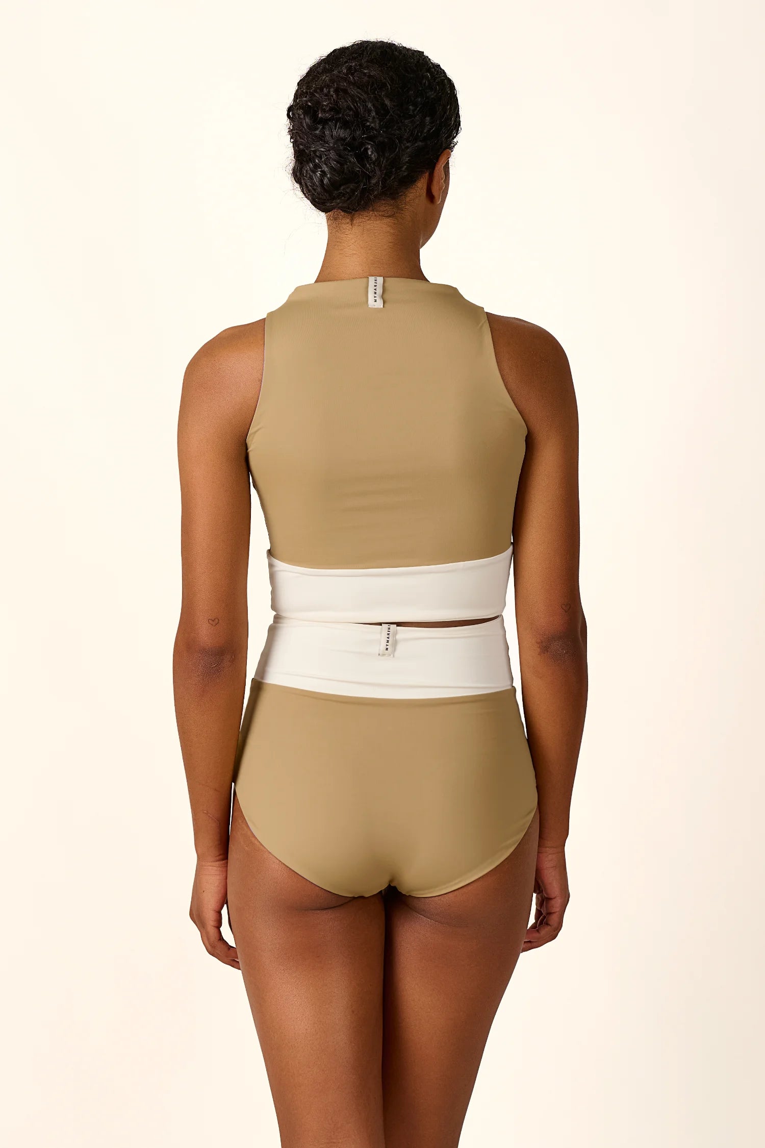 Rückansicht einer Frau in MYMARINI Badeanzug mit hohem Beinausschnitt und breiten Trägern in Beige und Weiß.#color_coast-desert