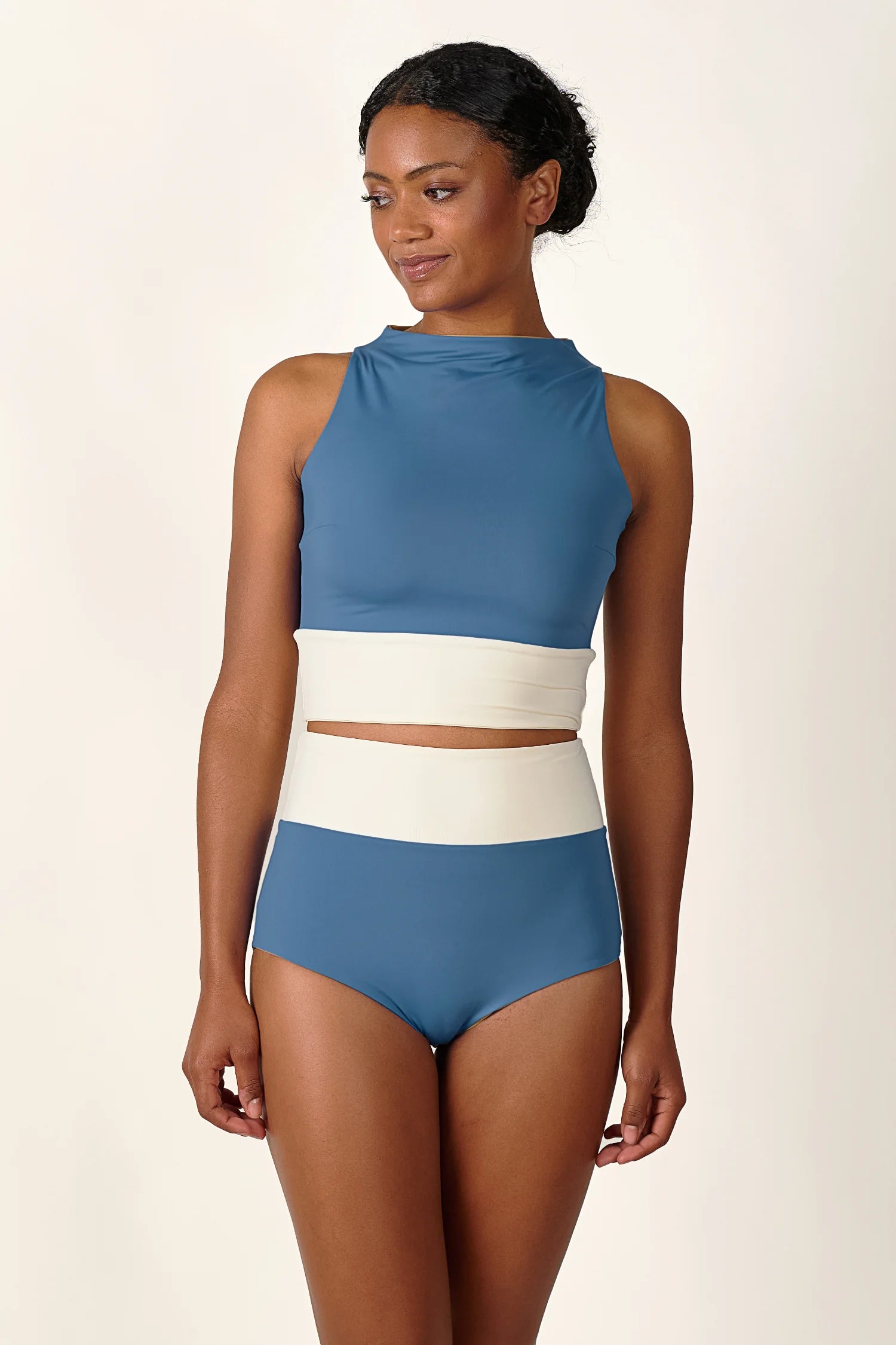 Frau trägt MYMARINI Badeanzug mit hochgeschlossenem Top und hoher Bikinihose in Blau und Weiß, lächelnd vor neutralem Hintergrund.#color_coast-desert