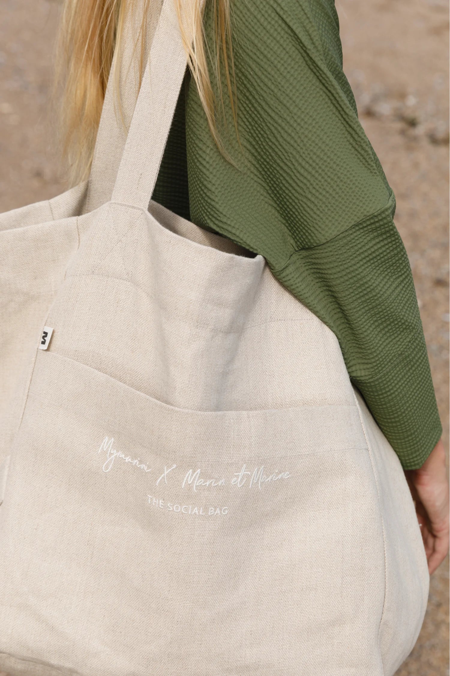 Person in grünem Oberteil trägt eine große, beige MYMARINI-Tasche mit Schriftzug.