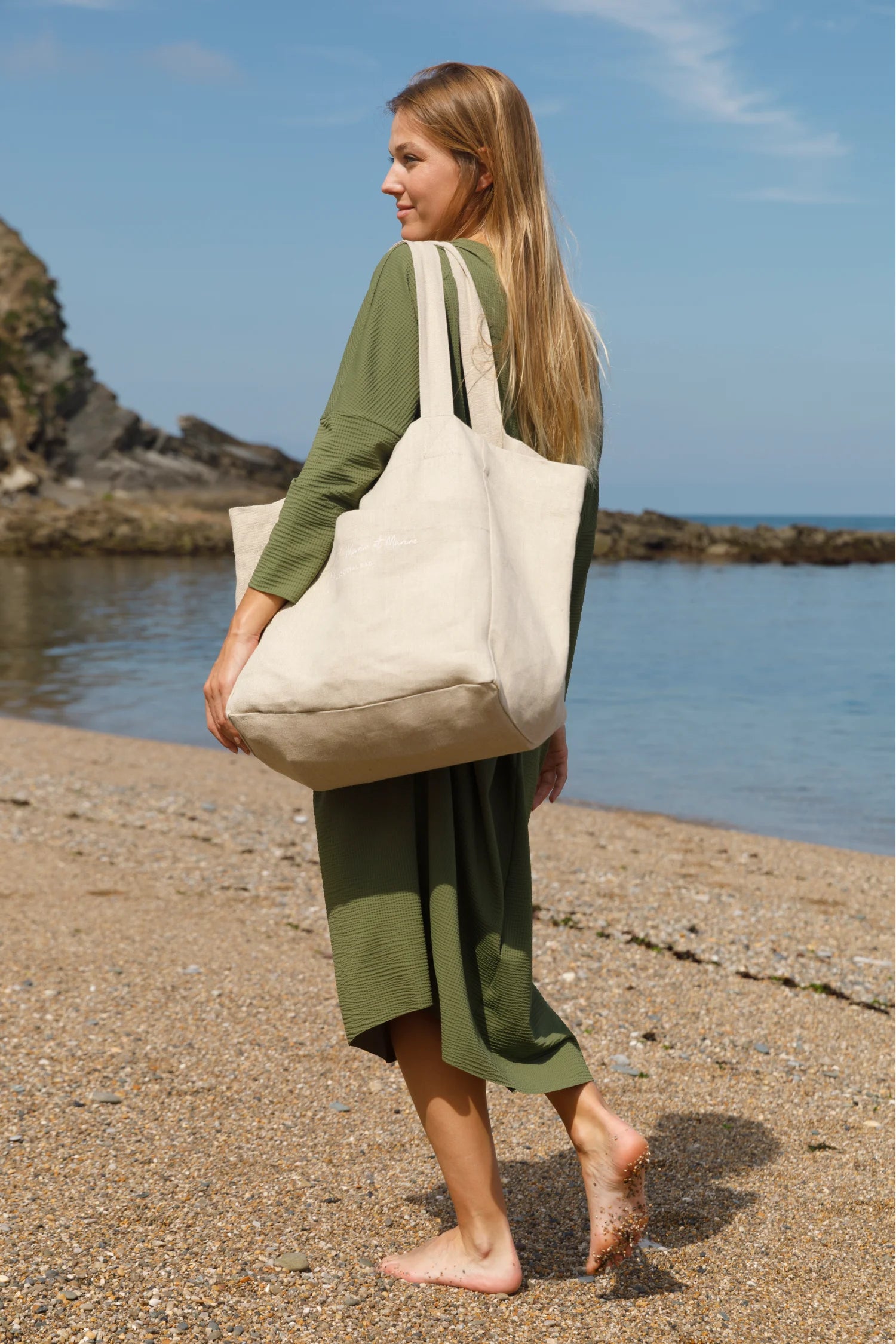 Frau am Strand in grünem MYMARINI-Kleid mit langen Ärmeln, trägt große beigefarbene Tasche.