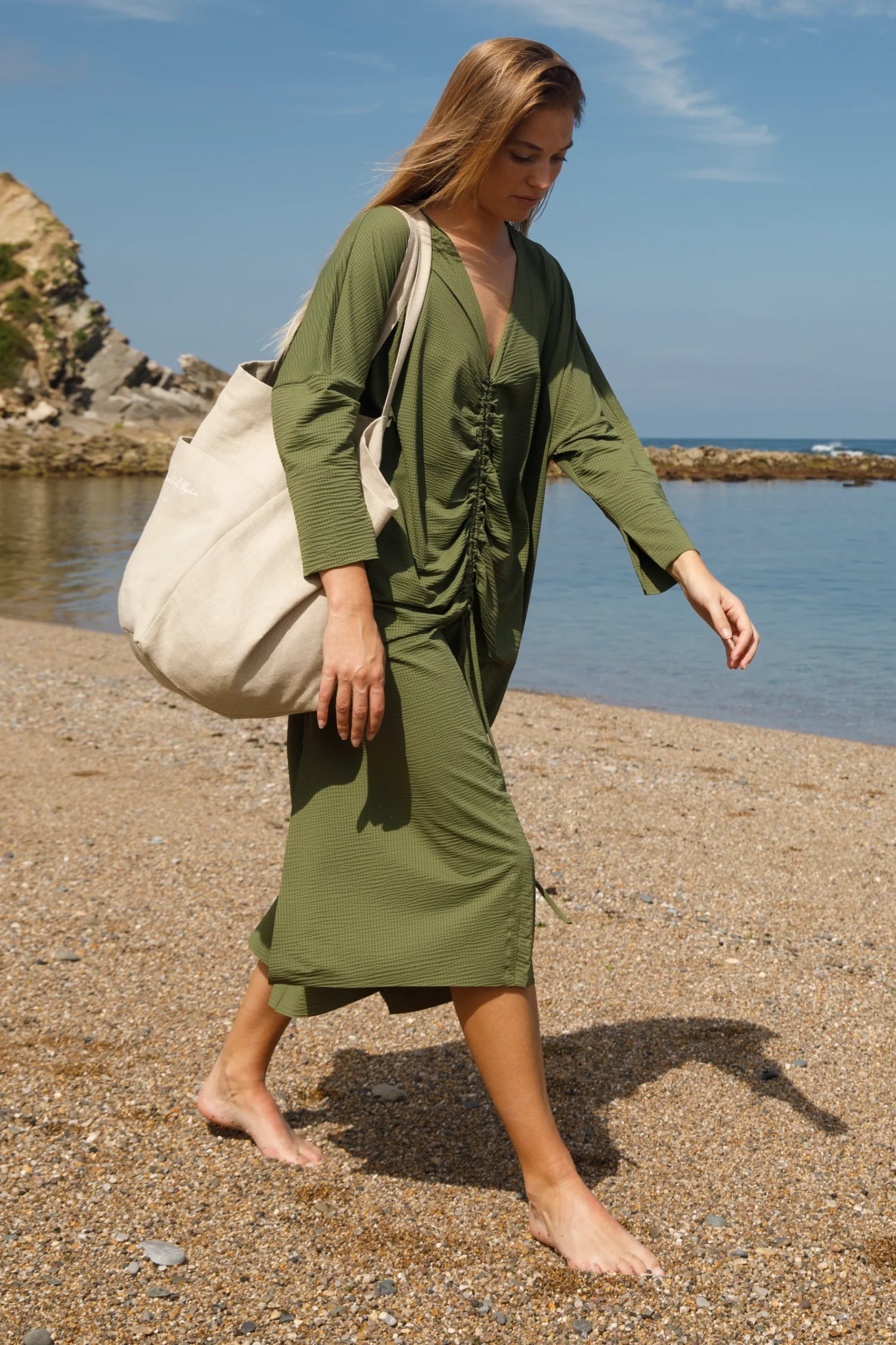 Frau am Strand in grünem MYMARINI-Kleid mit V-Ausschnitt, trägt große Stofftasche, barfuß, Meer und Felsen im Hintergrund.