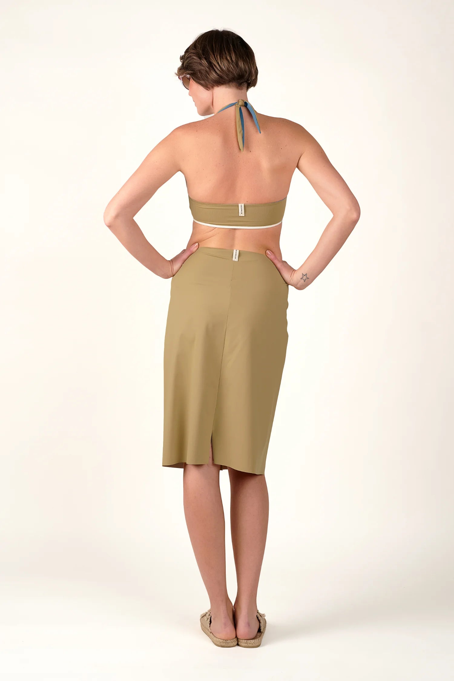 Frau in MYMARINI Neckholder-Bikinitop und Mid-rise Rock, Rückansicht, neutraler Hintergrund.#color_desert