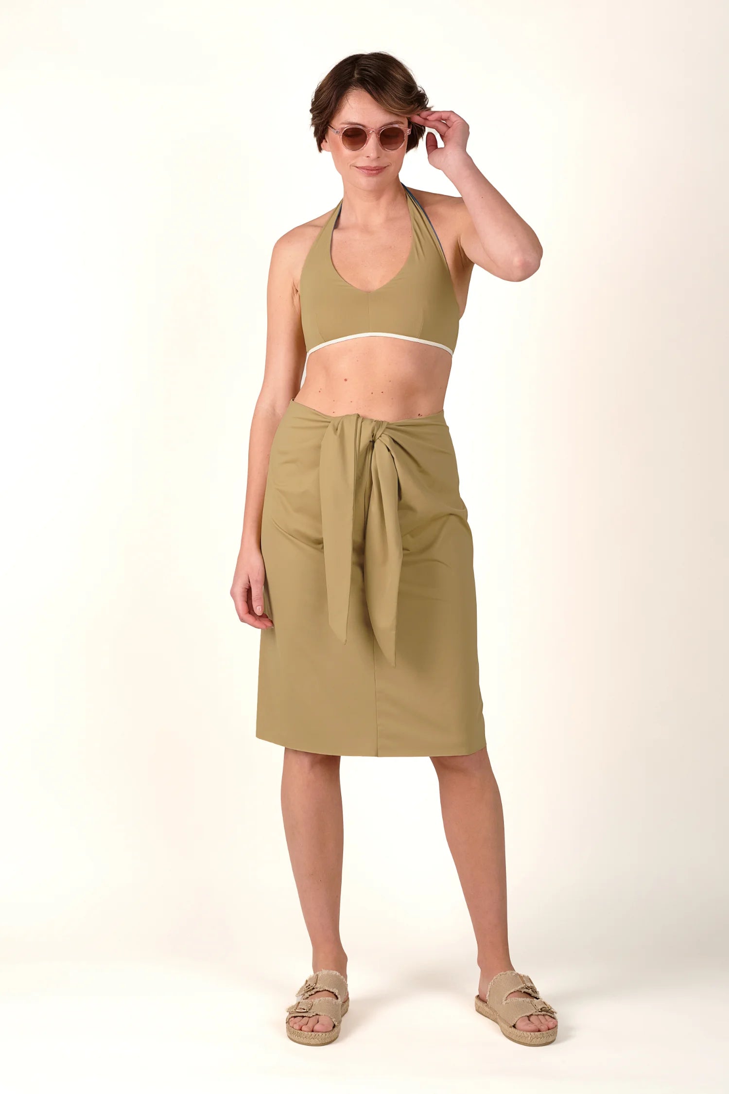 Frau in MYMARINI Neckholder-Bikinitop und Rock, beide in Beige. Sie trägt Sonnenbrille und Sandalen, steht vor weißem Hintergrund.#color_desert
