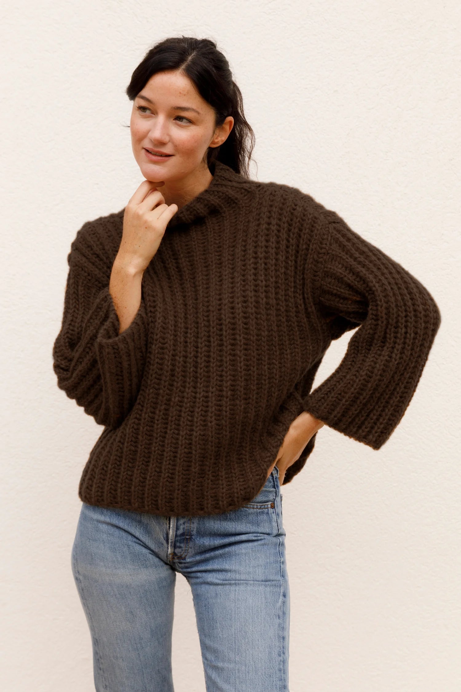 Frau in braunem Strickpullover und blauer Jeans vor hellem Hintergrund, lächelnd und mit einer Hand am Kinn.#color_chocolate