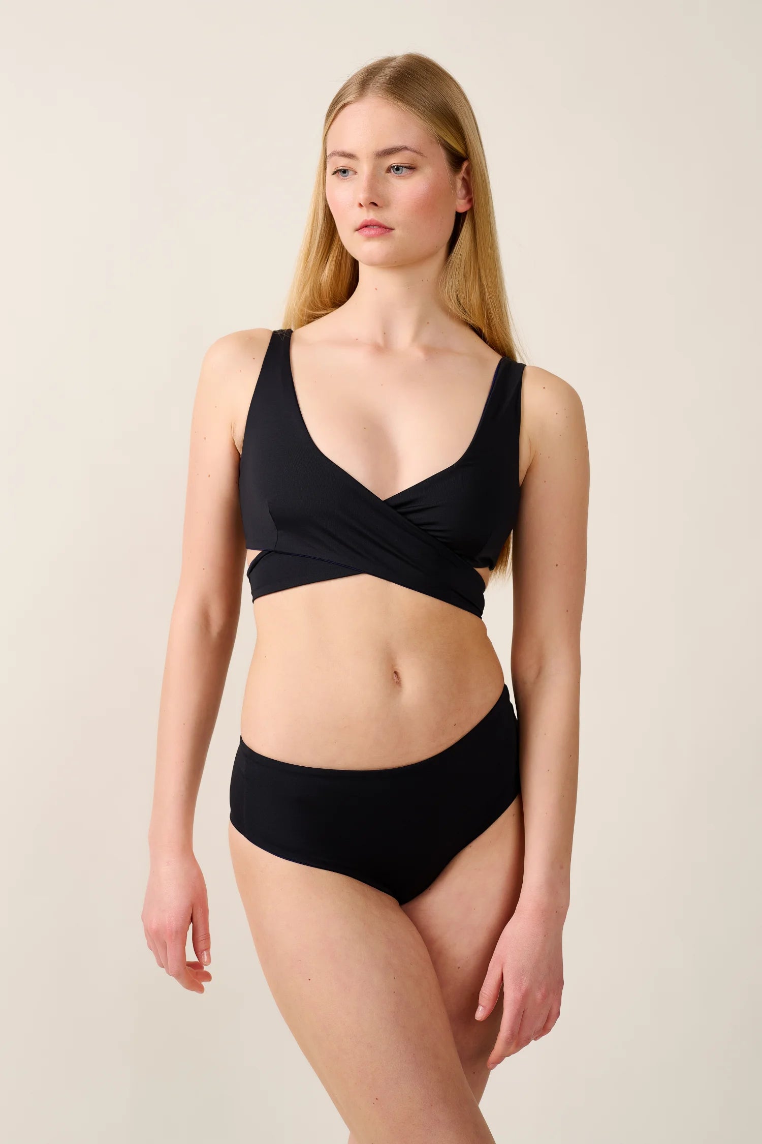 Frau trägt MYMARINI Bikinitop mit V-Ausschnitt und breite Träger, dazu passende Bikinihose, vor neutralem Hintergrund.#color_black-navy