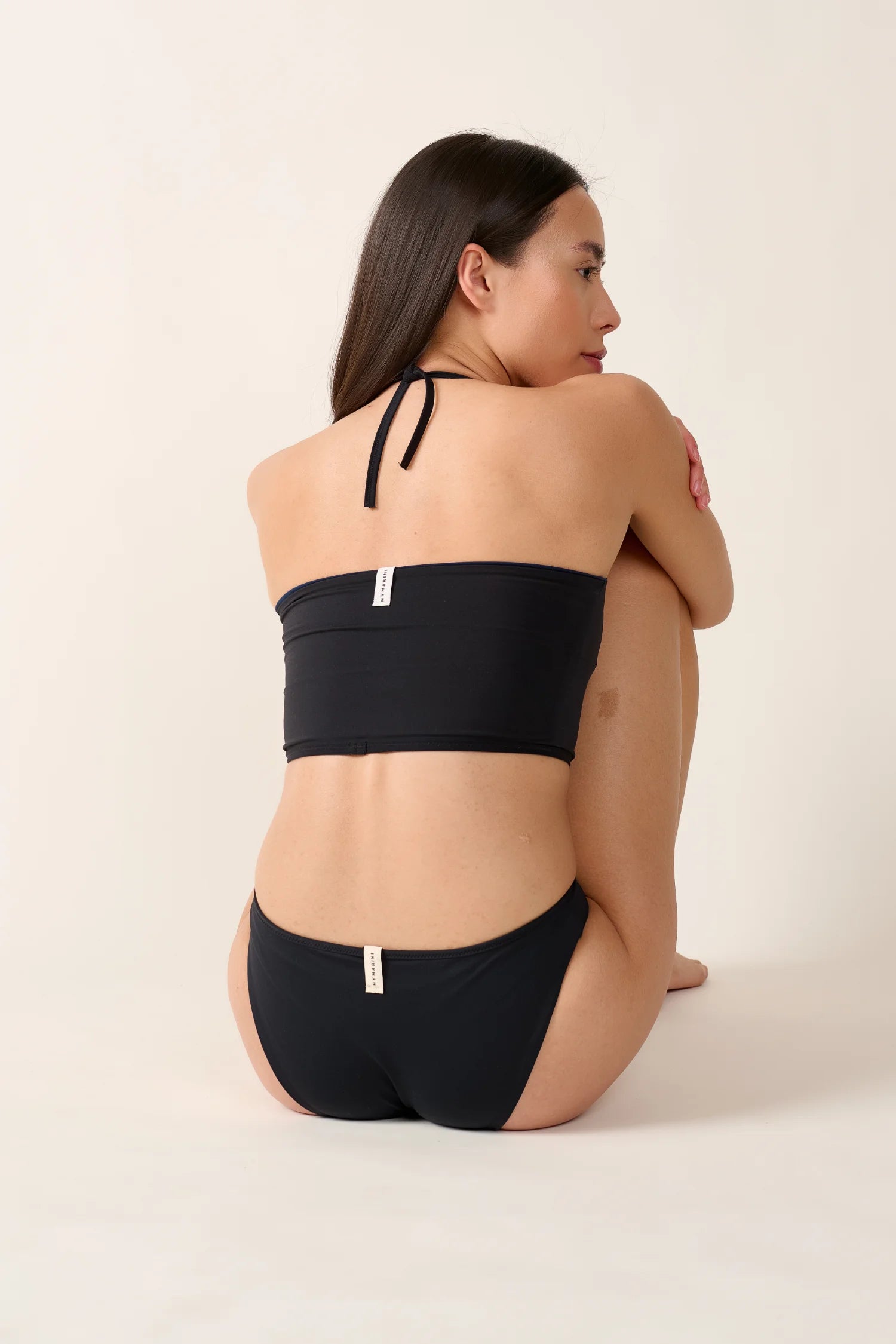 Frau in schwarzem MYMARINI Bikini mit Bandeau-Top und Neckholder, sitzt seitlich auf hellem Hintergrund.#color_black-navy
