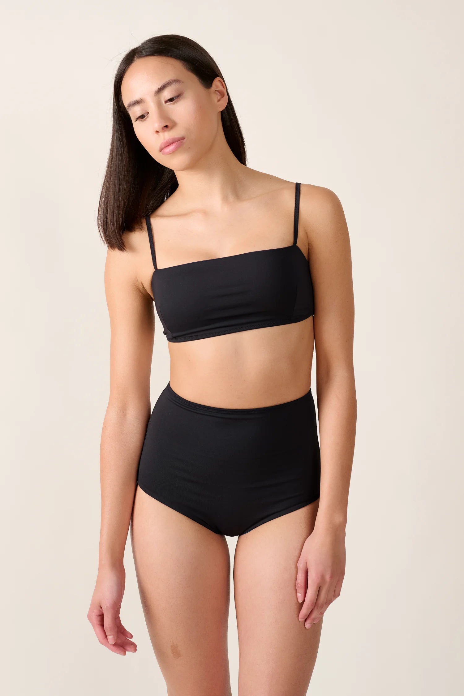 Frau trägt MYMARINI Bikinitop mit Spaghettiträgern und hoch geschnittene Bikinihose in Schwarz vor neutralem Hintergrund.#color_black-navy