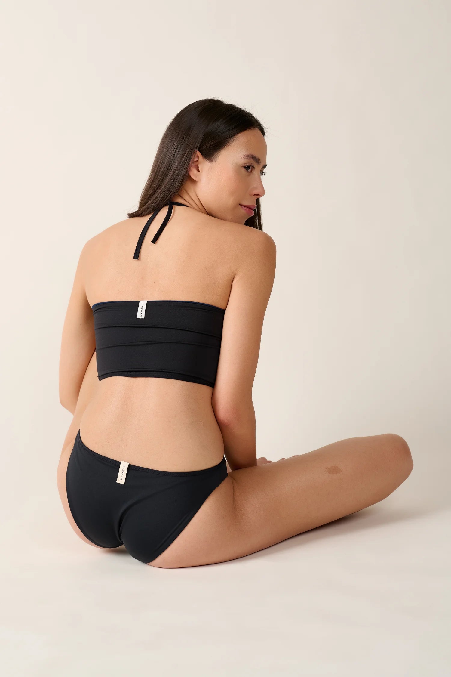 Frau in schwarzem MYMARINI-Bikini mit Bandeau-Top und Neckholder sitzt seitlich auf hellem Hintergrund.#color_black-navy