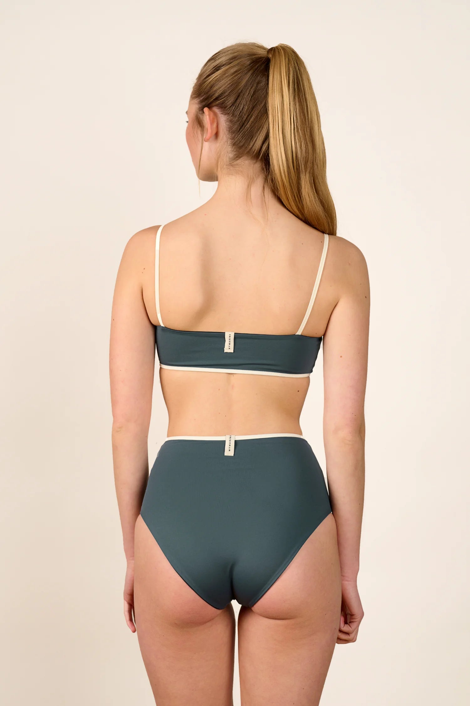 Frau trägt MYMARINI Bikini mit Bandeau-Top und High-Waist-Hose in Grün, Rückansicht, vor neutralem Hintergrund.#color_black-grey-white