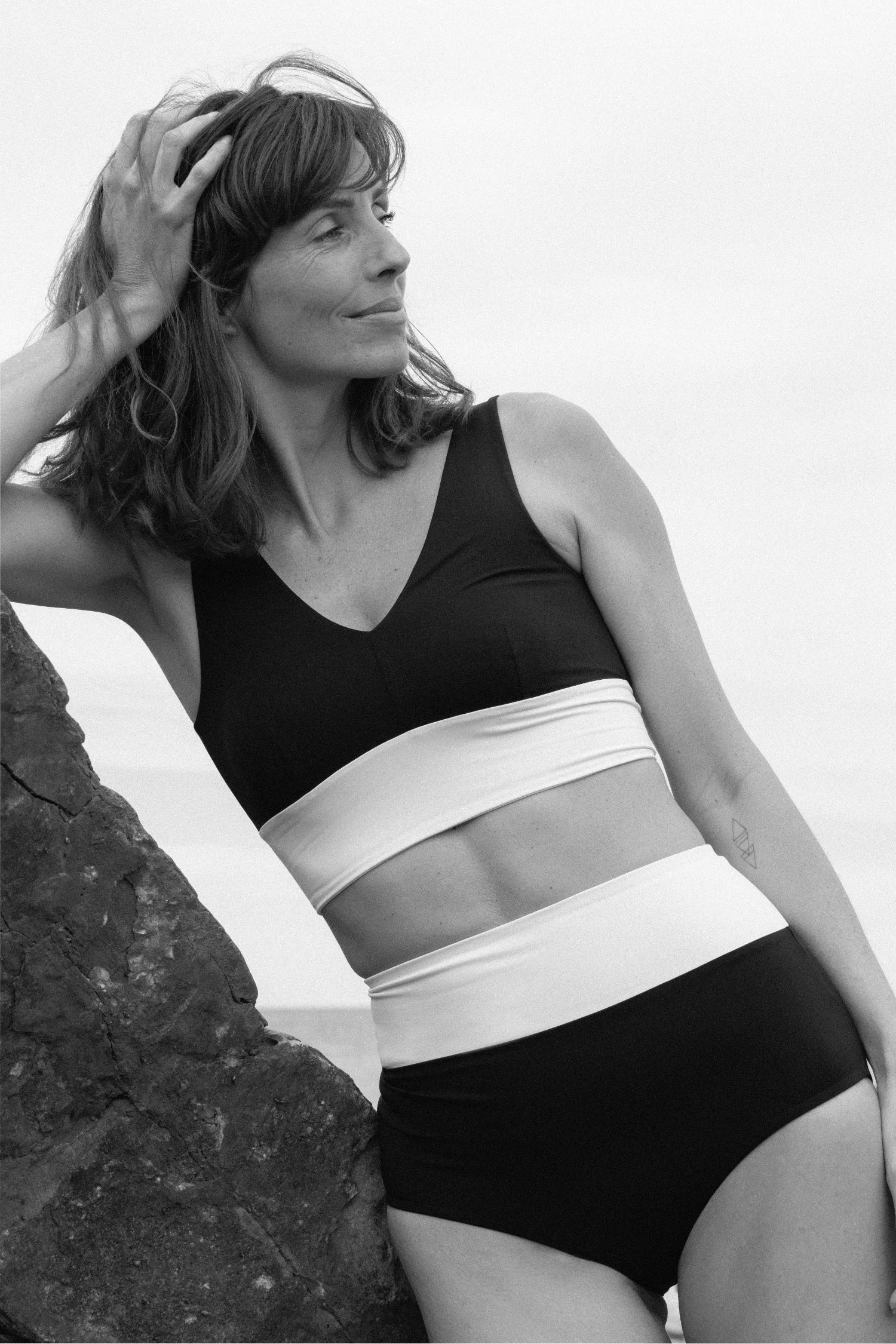 Frau in MYMARINI Bikini mit V-Ausschnitt und High Waist, lehnt an Felsen, schaut lächelnd in die Ferne.#color_black-grey-white