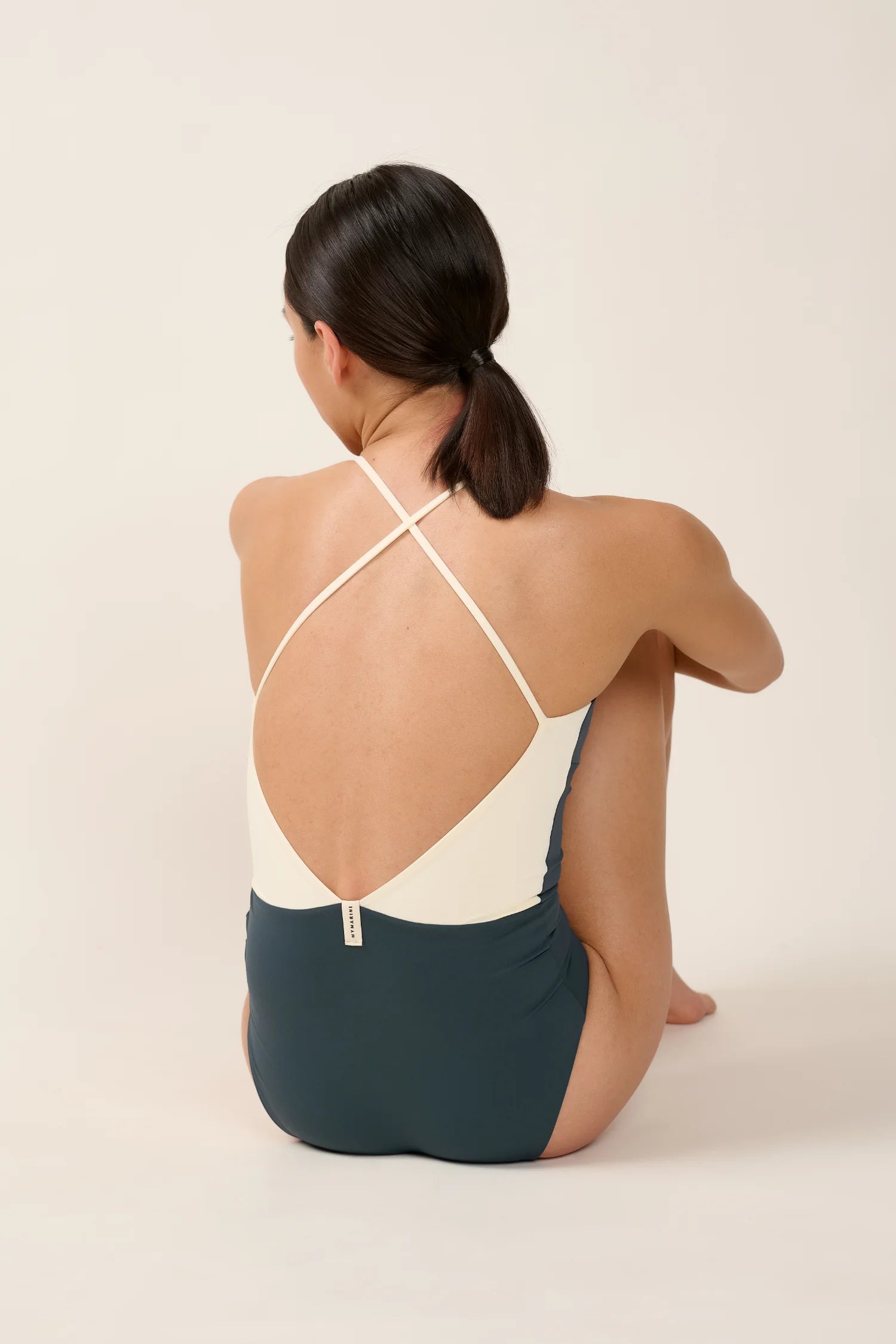 Frau sitzt im MYMARINI Badeanzug mit tiefem Rückenausschnitt und Spaghettiträgern auf hellem Hintergrund.#color_black-grey-white