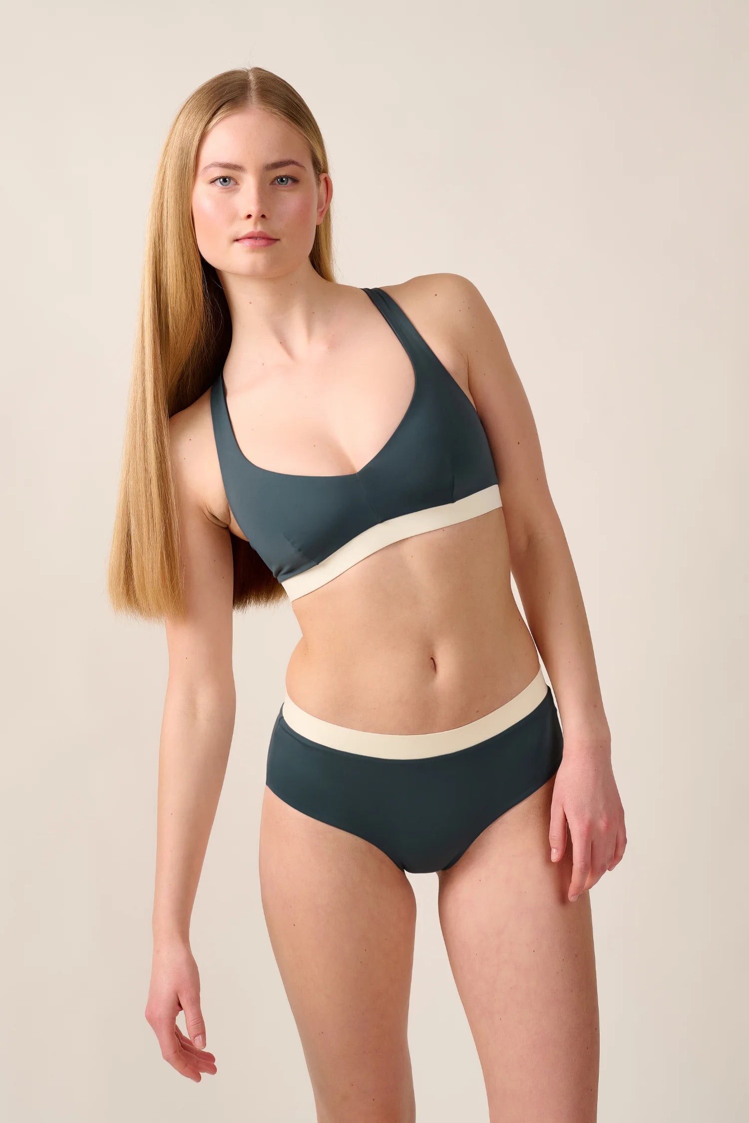 Frau trägt MYMARINI Bikini mit breiten Trägern und hohem Beinausschnitt in Dunkelgrün und Creme vor neutralem Hintergrund.#color_black-grey-white