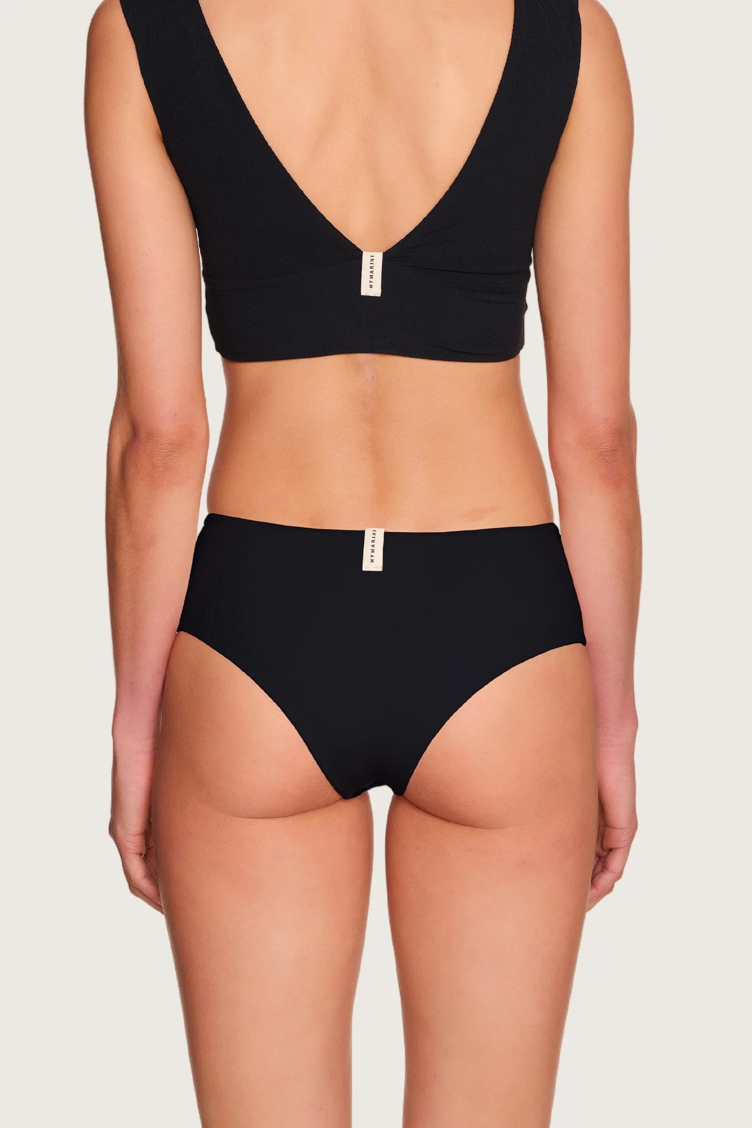 Rückansicht einer Person in schwarzem MYMARINI-Bikinitop mit V-Ausschnitt und passender Bikinihose, mid rise.#color_black