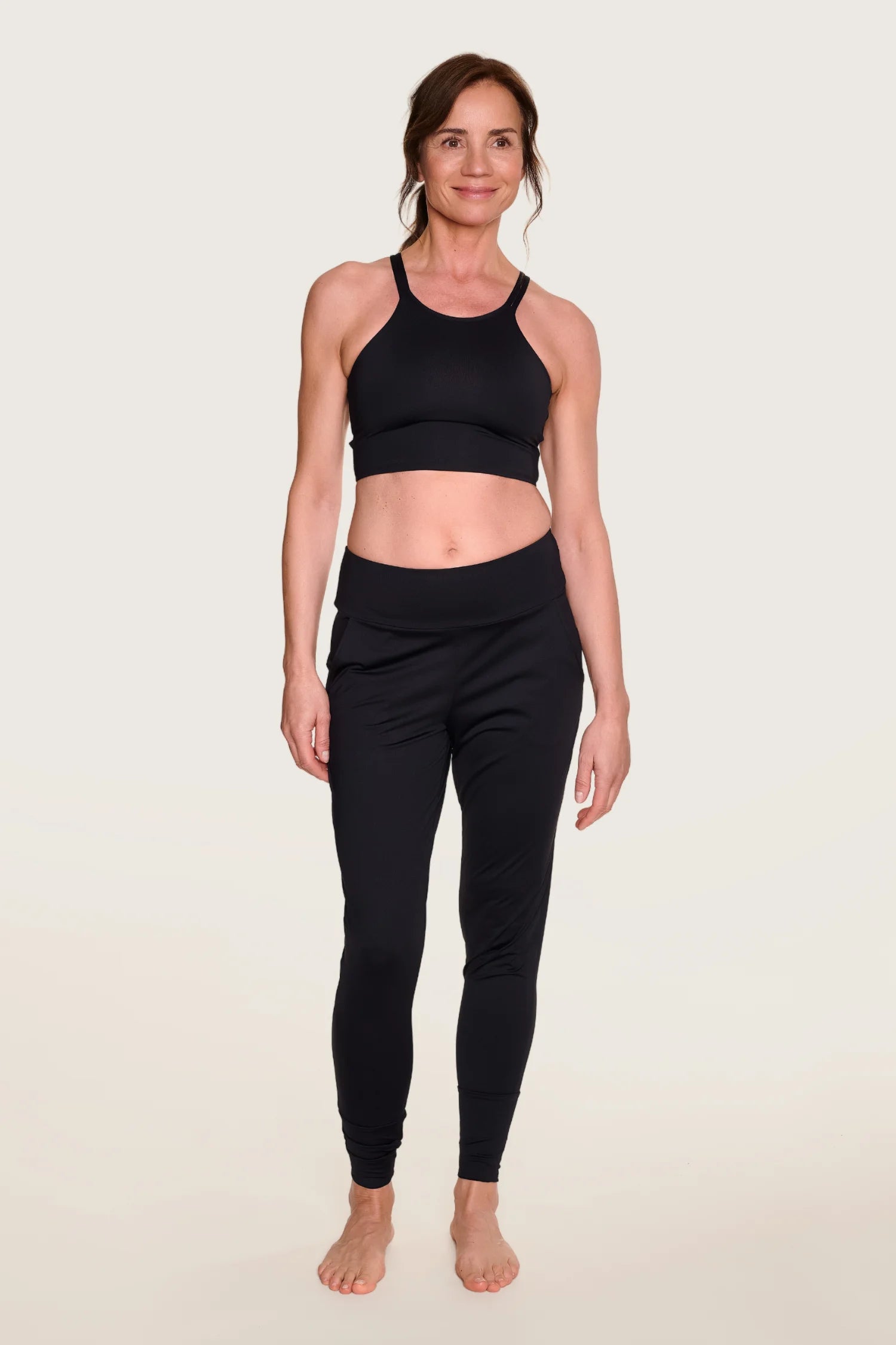 Frau trägt MYMARINI Sport-BH mit breiten Trägern und Leggings, beide in Schwarz, vor hellem Hintergrund.#color_black