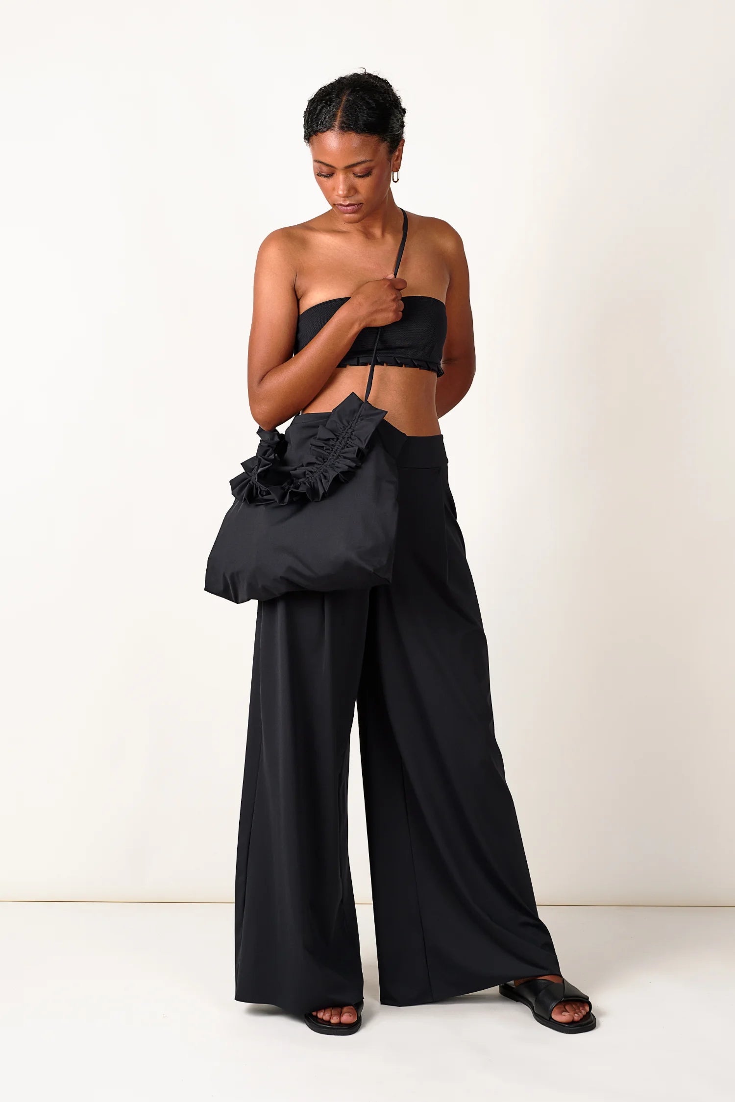 Frau in schwarzem MYMARINI Bandeau-Bikinitop und weiter Hose, hält schwarze Tasche mit Rüschen.#color_black