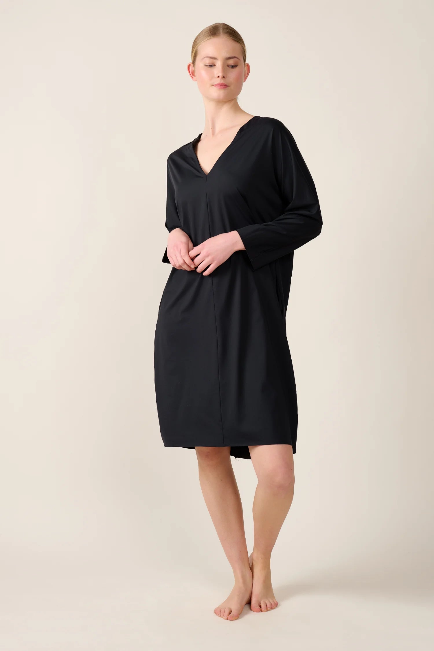 Person trägt ein schwarzes MYMARINI-Kleid mit V-Ausschnitt. Sie steht barfuß vor neutralem Hintergrund.#color_black