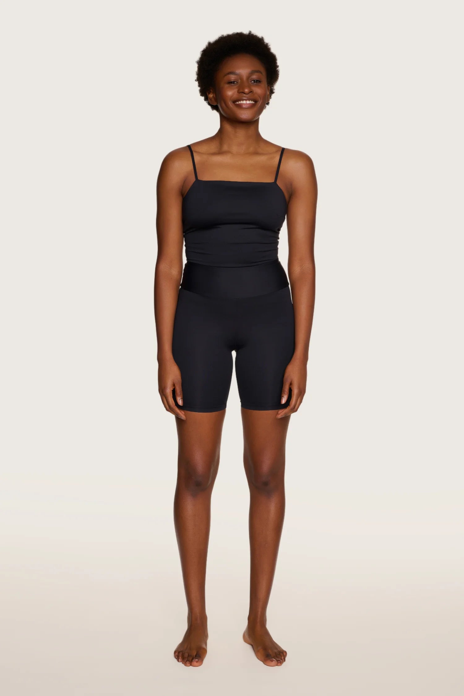 Frau trägt MYMARINI Top mit Spaghettiträgern und High-Waist-Shorts, lächelnd vor neutralem Hintergrund.#color_black