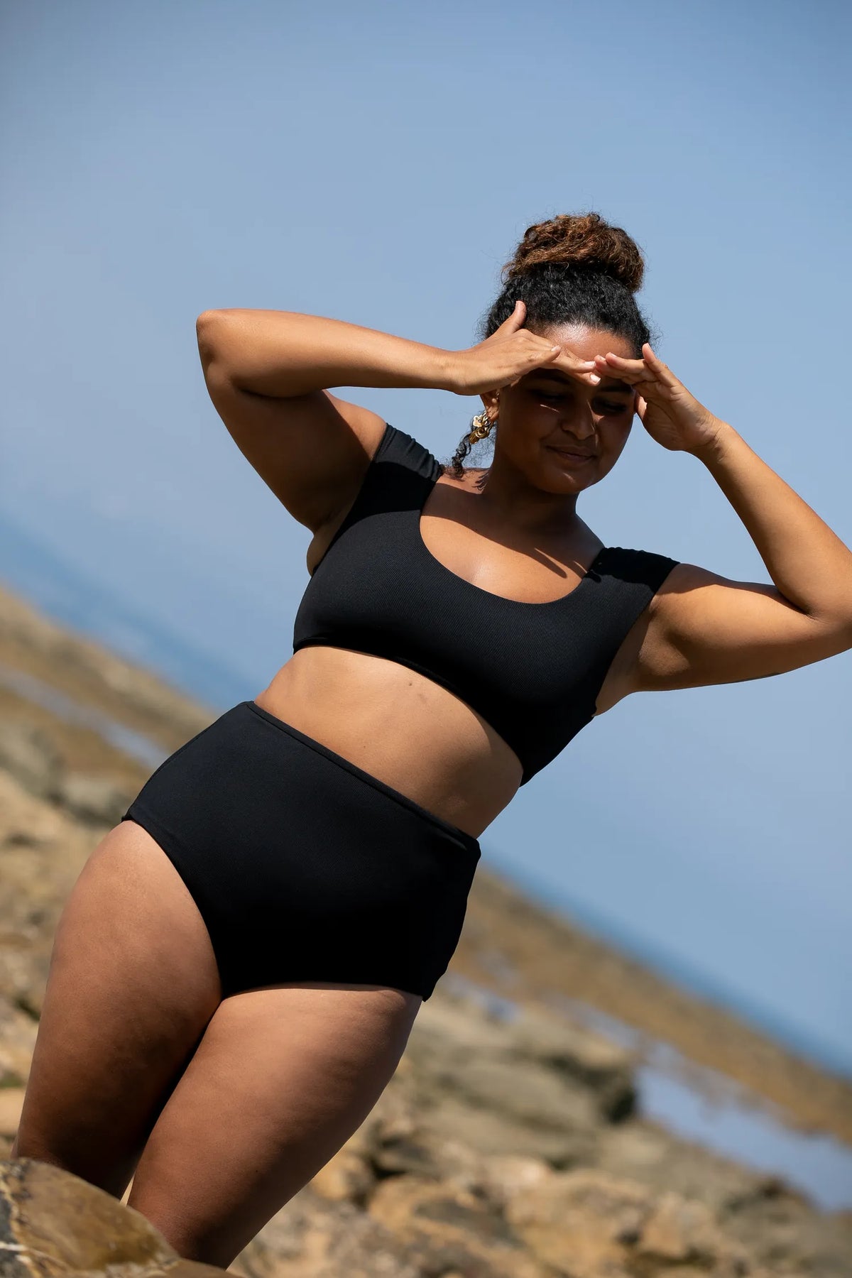 Woman on a rocky shore, hands at her forehead, in the black MYMARINI SHOULDER TOP bikini top#color_black-ribbed