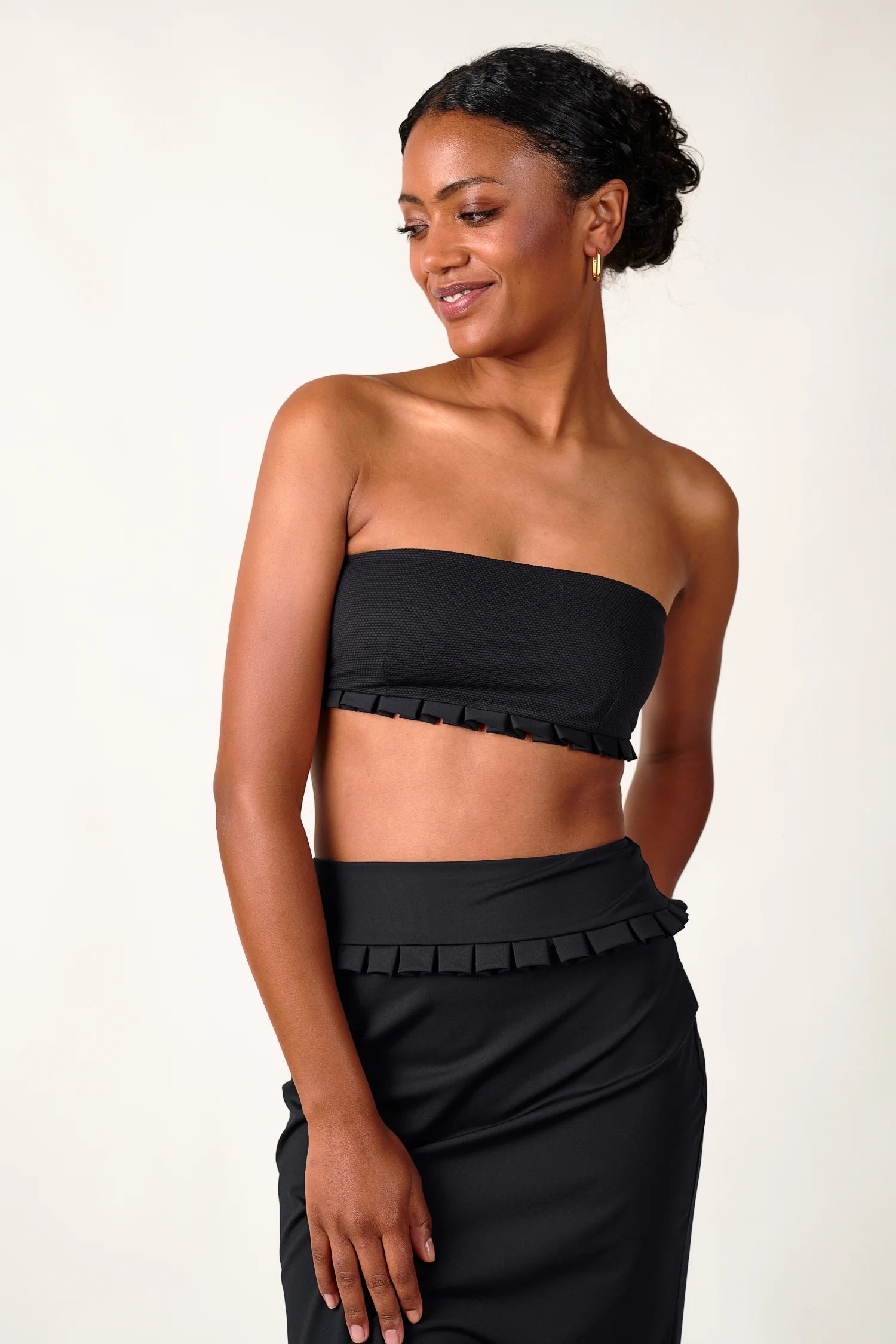 Frau trägt MYMARINI Bandeau-Bikinitop und Rock in Schwarz, lächelnd, mit dezentem Rüschenbesatz, vor hellem Hintergrund.#color_black