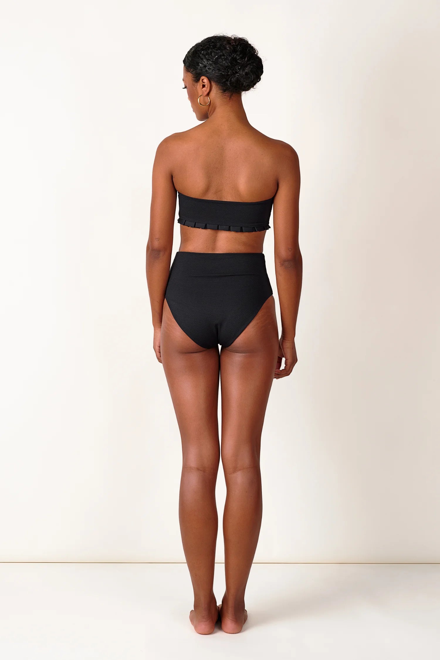 Person von hinten in schwarzem MYMARINI Bandeau-Bikinitop und High-Waist-Bikinihose vor hellem Hintergrund.#color_black