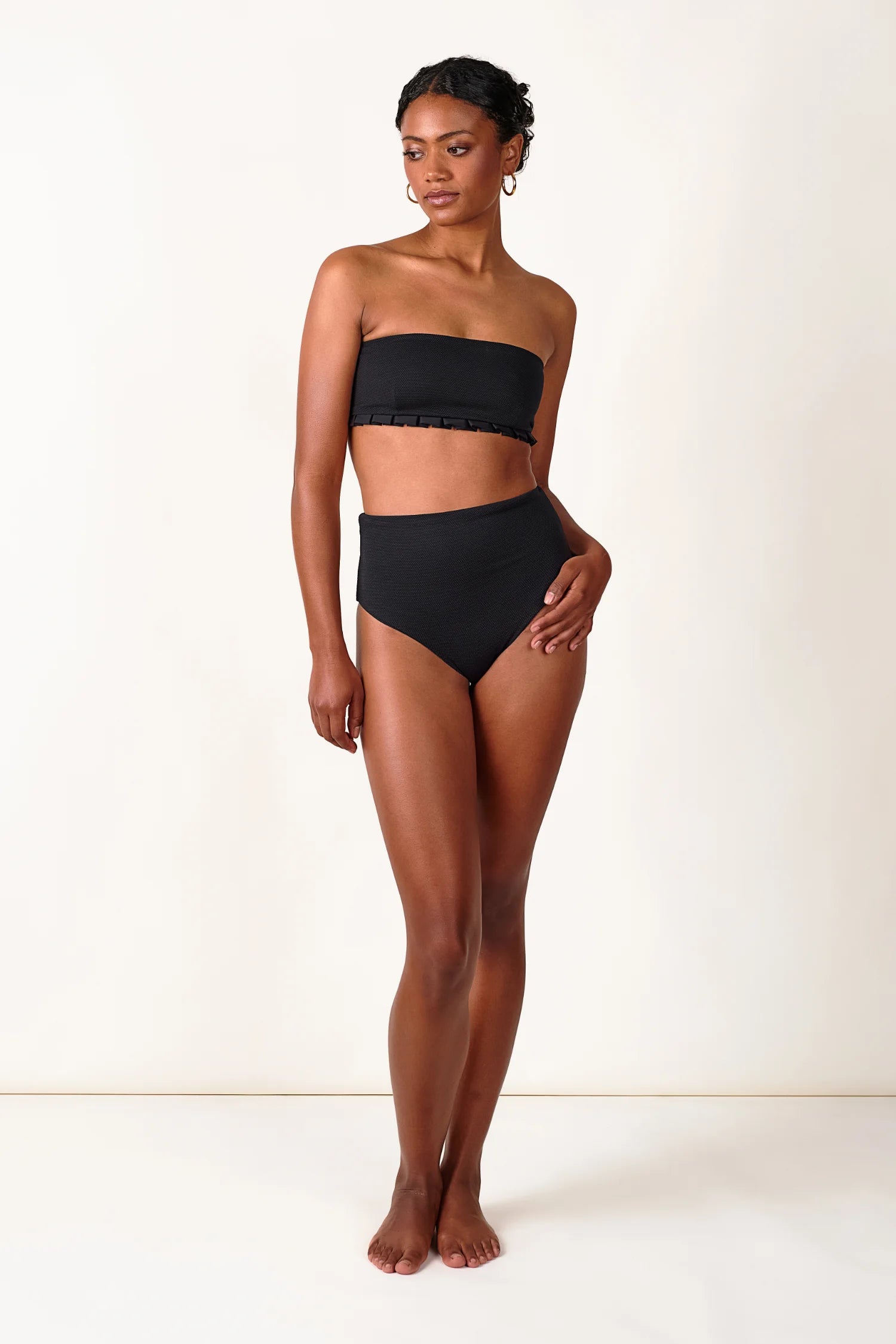 Frau trägt schwarzen MYMARINI Bandeau-Bikinitop und High-Waist-Bikinihose vor neutralem Hintergrund.#color_black