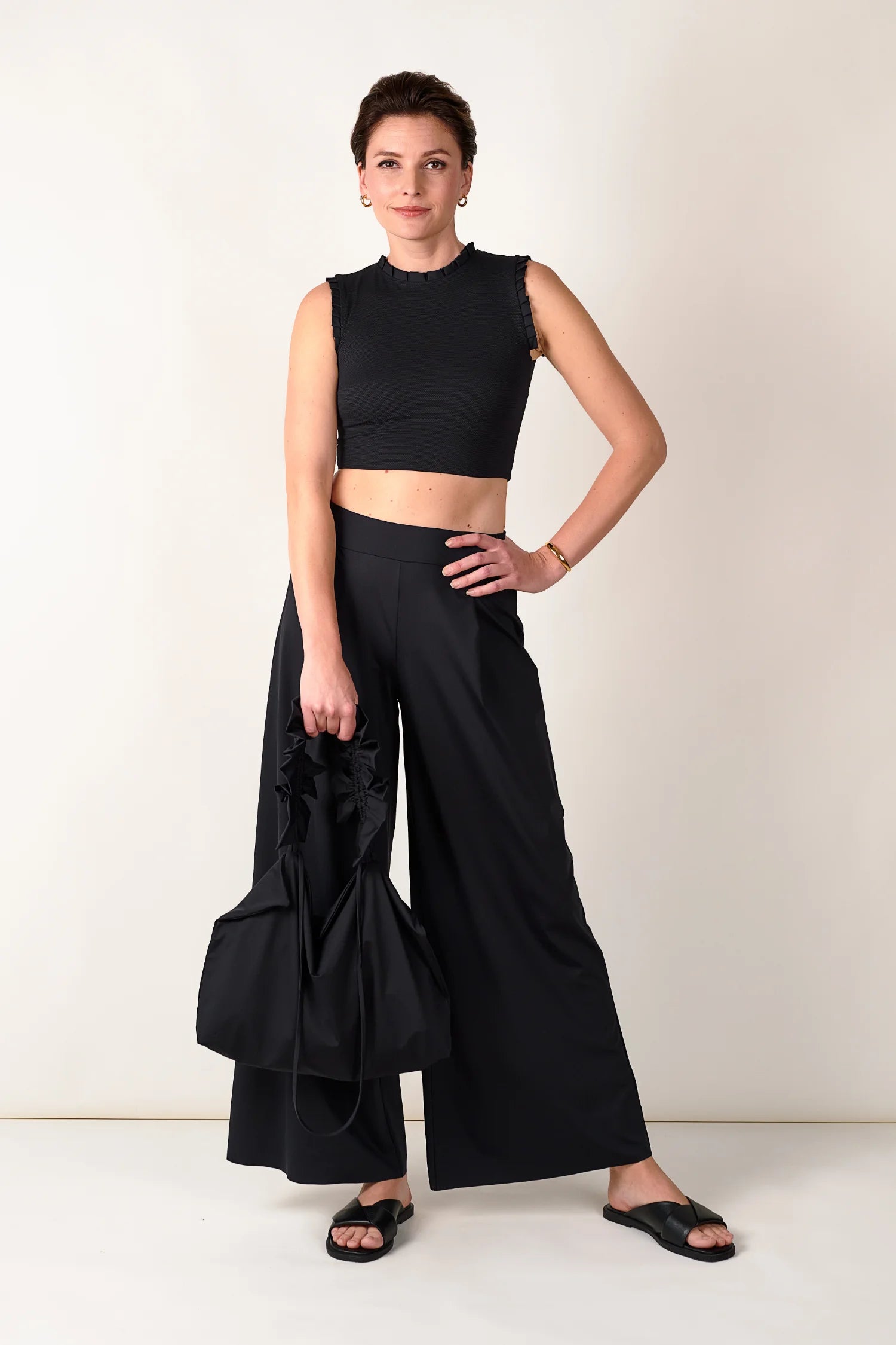 Frau in schwarzem MYMARINI-Top mit breiten Trägern und schwarzer Hose, hält schwarze Tasche, vor hellem Hintergrund.#color_black