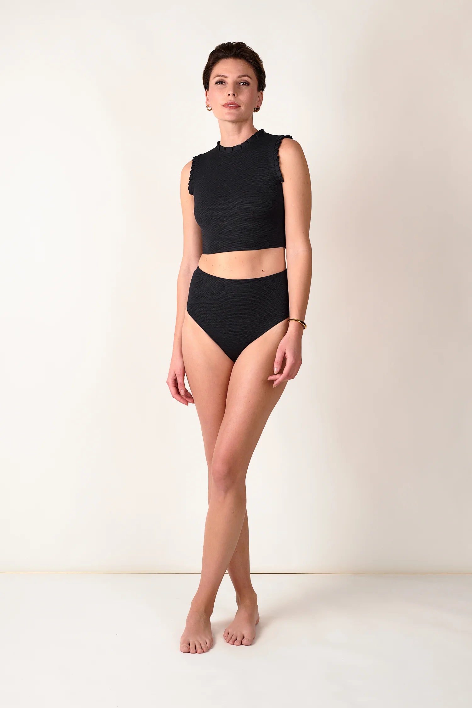 Frau trägt MYMARINI Bikinihose mit hohem Beinausschnitt und hochgeschlossenem Top in Schwarz vor neutralem Hintergrund.#color_black