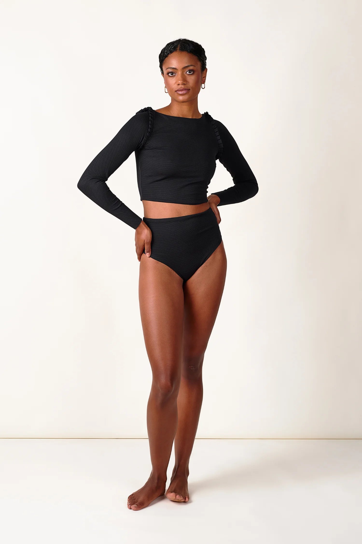Frau trägt MYMARINI Bikinihose mit hohem Beinausschnitt und langärmliges Top in Schwarz, stehend vor neutralem Hintergrund.#color_black