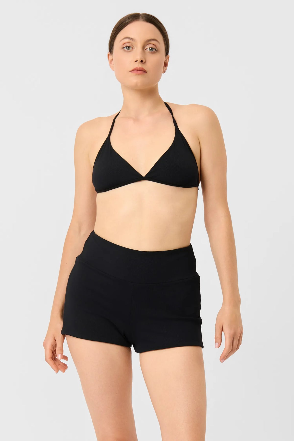 Woman in the black MYMARINI TRIANGLE TOP bikini top worn as a halterneck, with black shorts, front view#color_black-ribbed