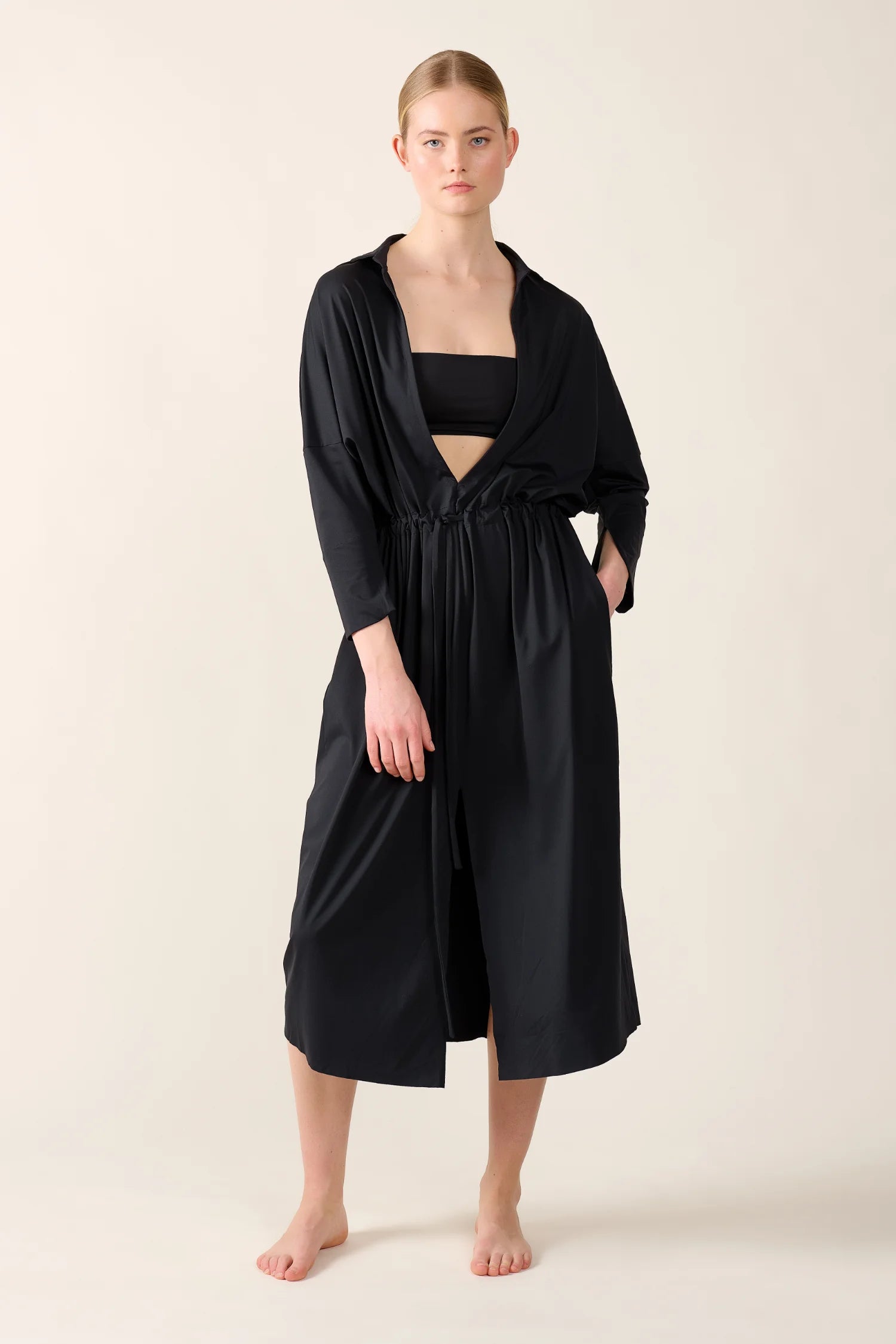 Person trägt ein schwarzes MYMARINI Bikinitop mit Bandeau-Schnitt und ein schwarzes, langes Kleid mit langen Ärmeln.#color_black